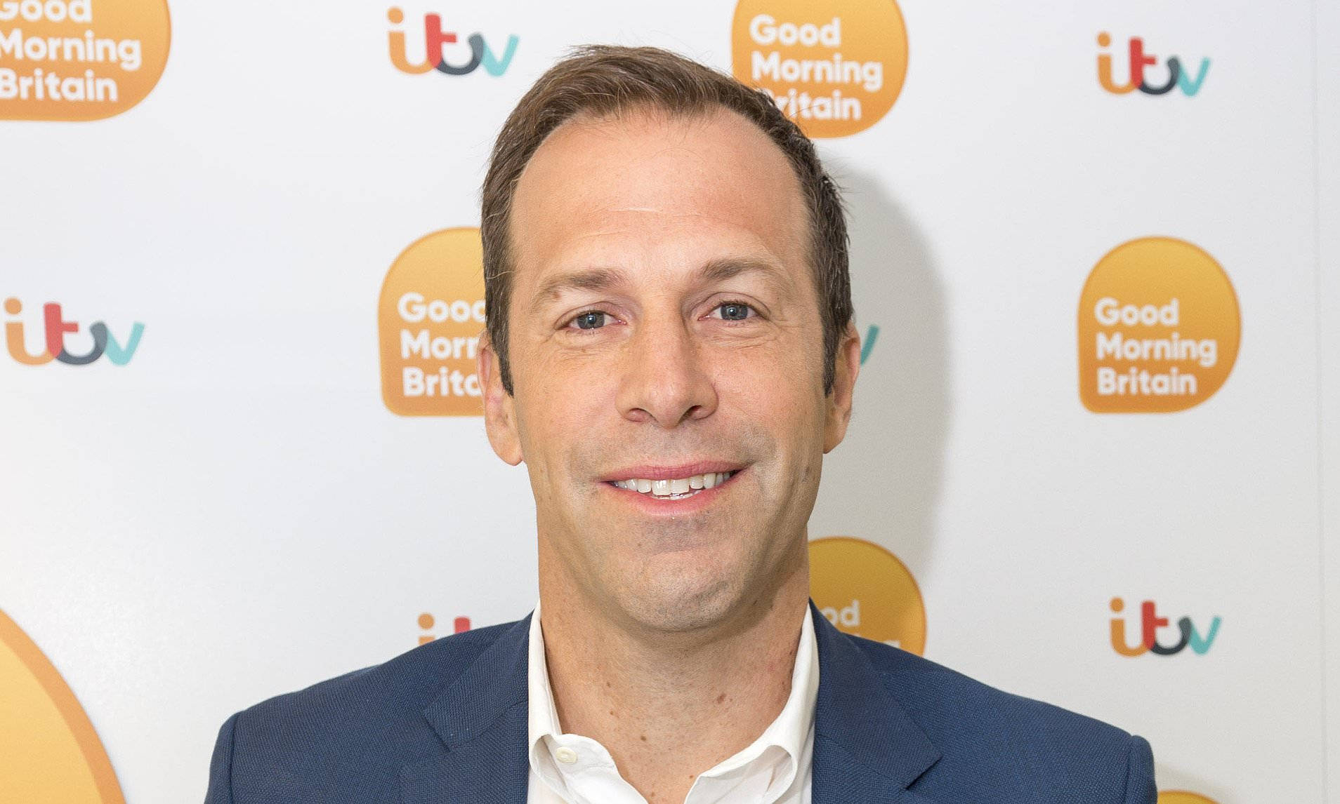 運動 桌布 壁紙 — 一位微笑的男士身穿藍色西裝，站在帶有「Good Morning Britain」標誌的品牌背景前。
