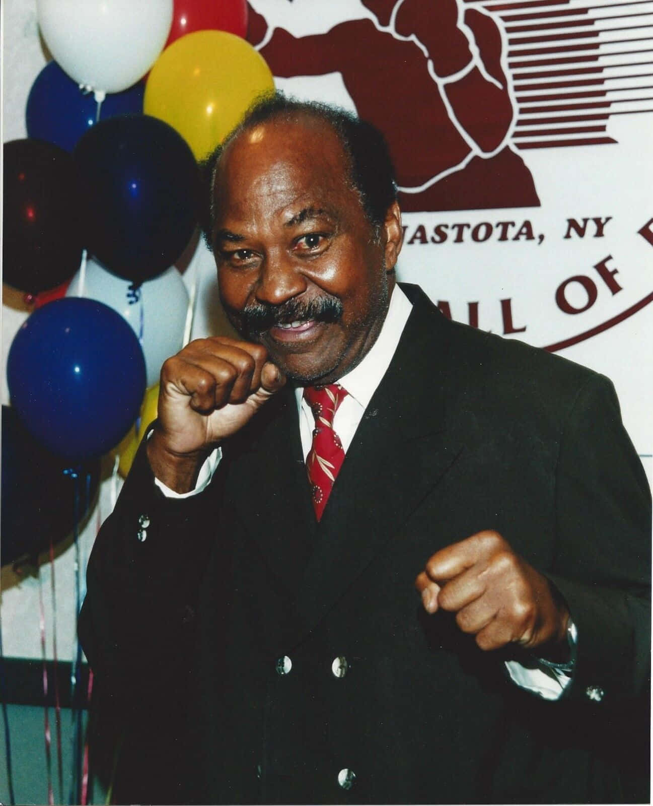 boxer 桌布 壁紙 — 一位微笑的男士身穿西裝和紅色領帶，在彩色氣球和「Hall of Fame」橫幅前擺出拳擊姿勢。