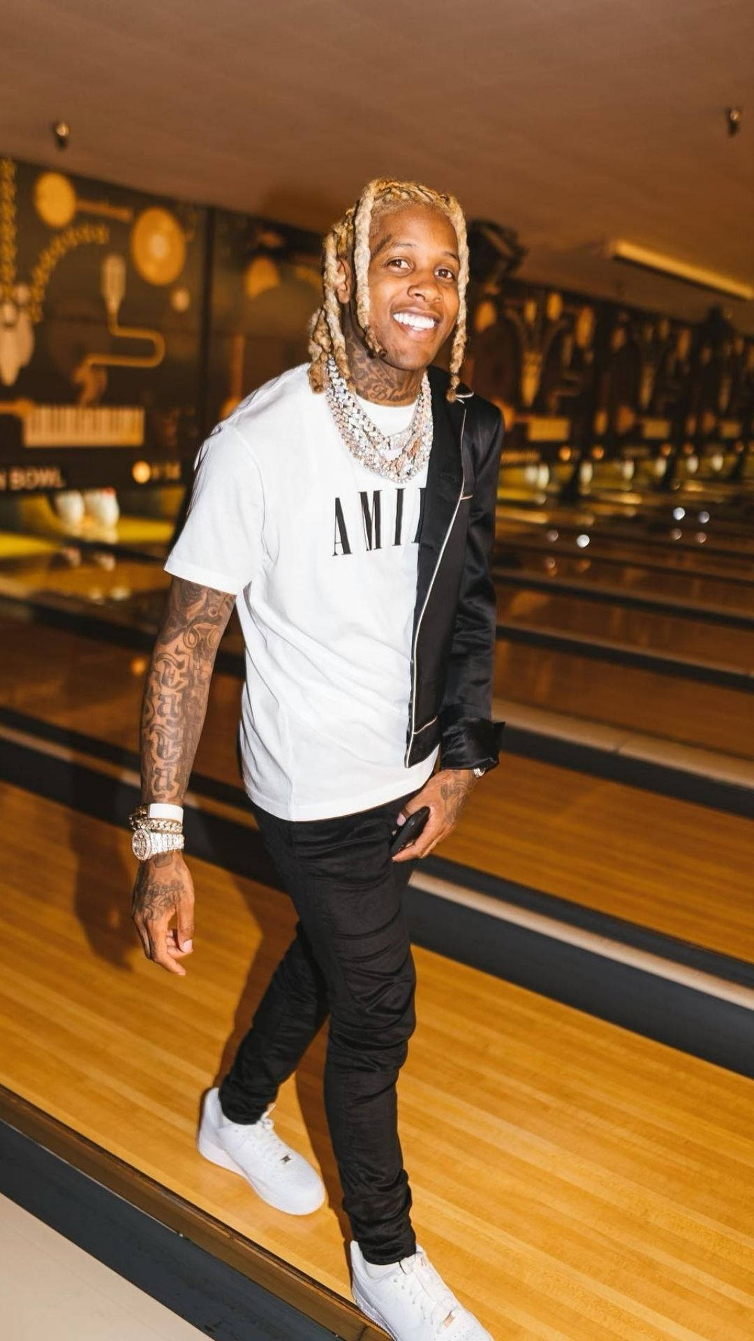 Lil Durk 桌布 壁紙 — 一位微笑的男子，梳著辮子頭，身上有刺青，自信地走在拋光的木製保齡球道上，身穿白色T恤、黑色外套和白色運動鞋。