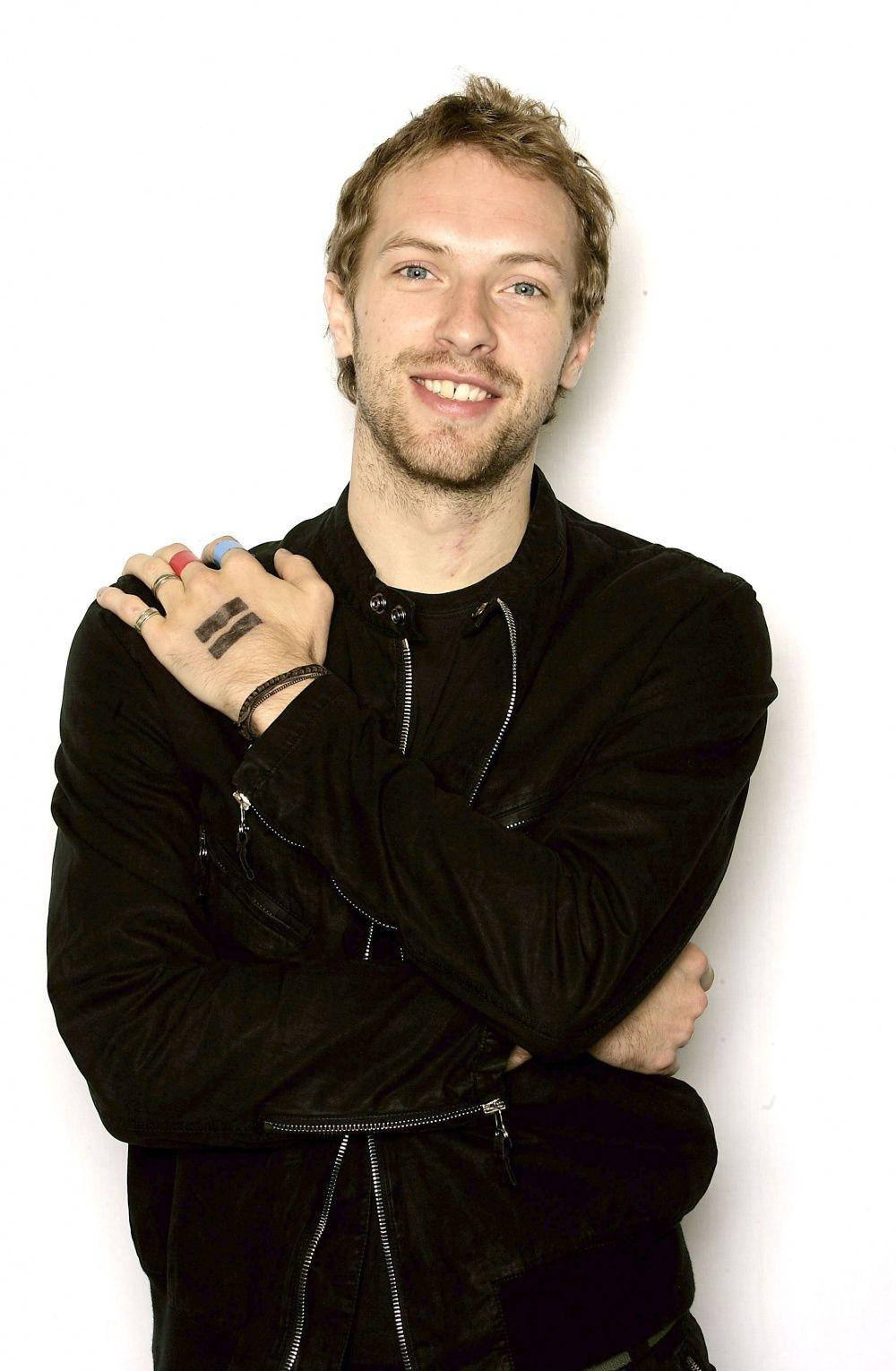 Chris Martin 桌布 壁紙 — 一位微笑的男子，淺色頭髮和鬍鬚，穿著黑色拉鍊夾克，背景為純白色。