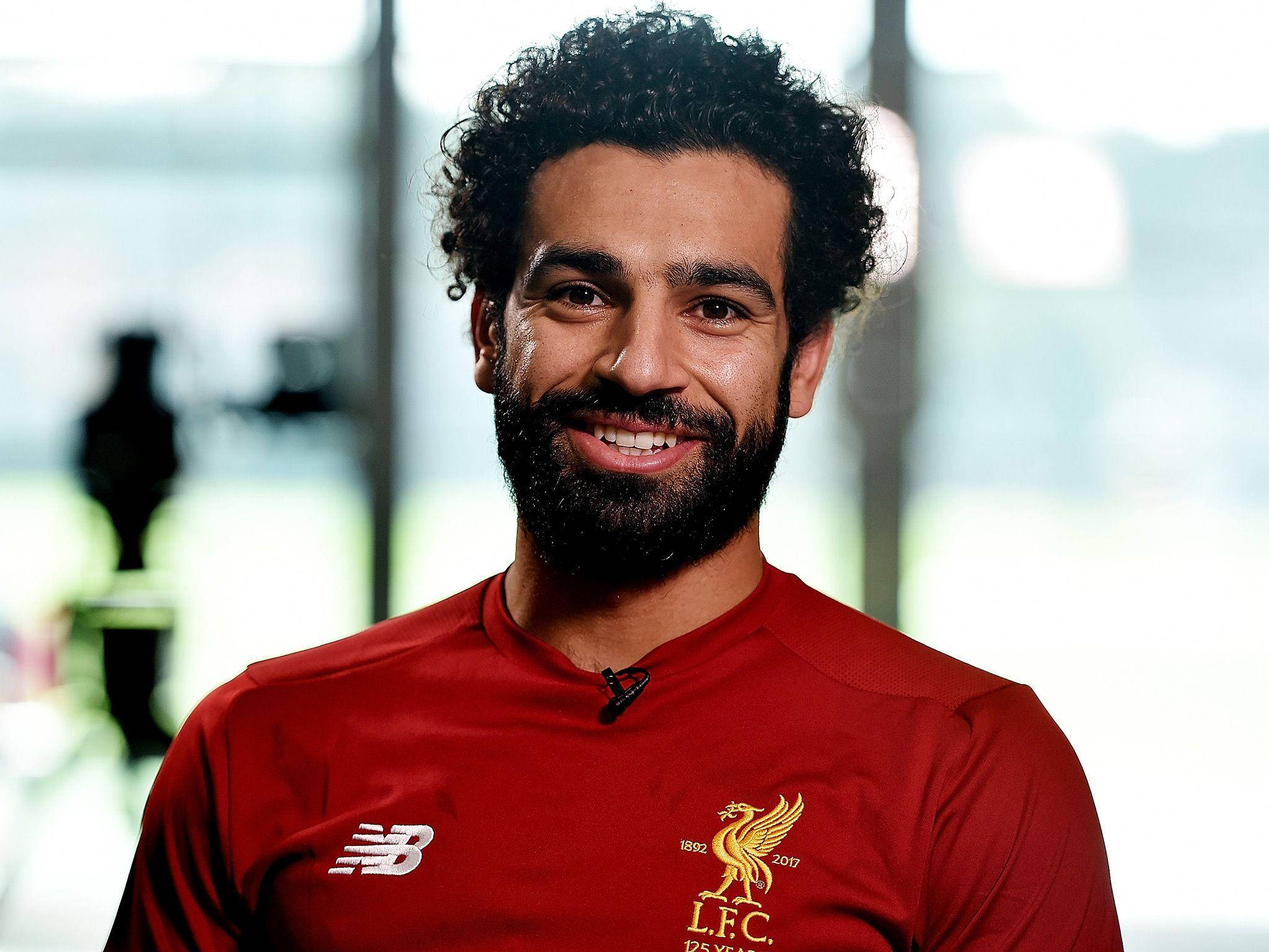Salah 桌布 壁紙 — 一位微笑的男子，留著捲曲深色頭髮和鬍鬚，身穿紅色利物浦足球俱樂部球衣，上有金色會徽和New Balance標誌。