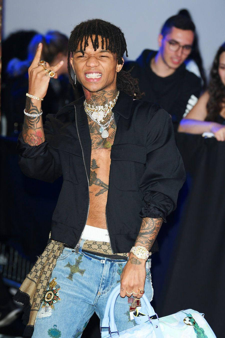 Swae Lee 桌布 壁紙 — 一位微笑的男子，留著辮子頭和刺青，穿著黑色外套和圖案牛仔褲，比出和平手勢。