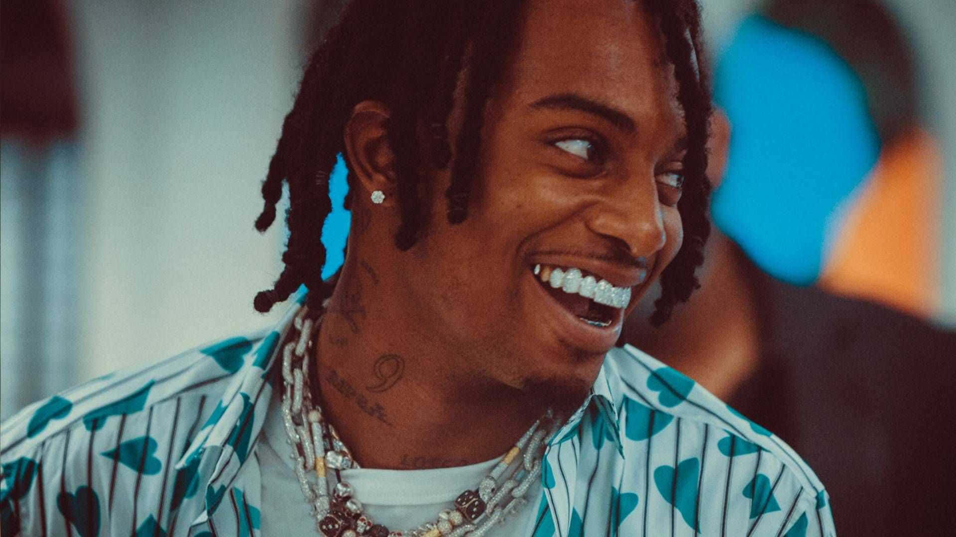 Playboi Carti 桌布 壁紙 — 一位微笑的男子，留著辮子頭和鑽石牙齒，穿著心形圖案襯衫和層疊項鍊。