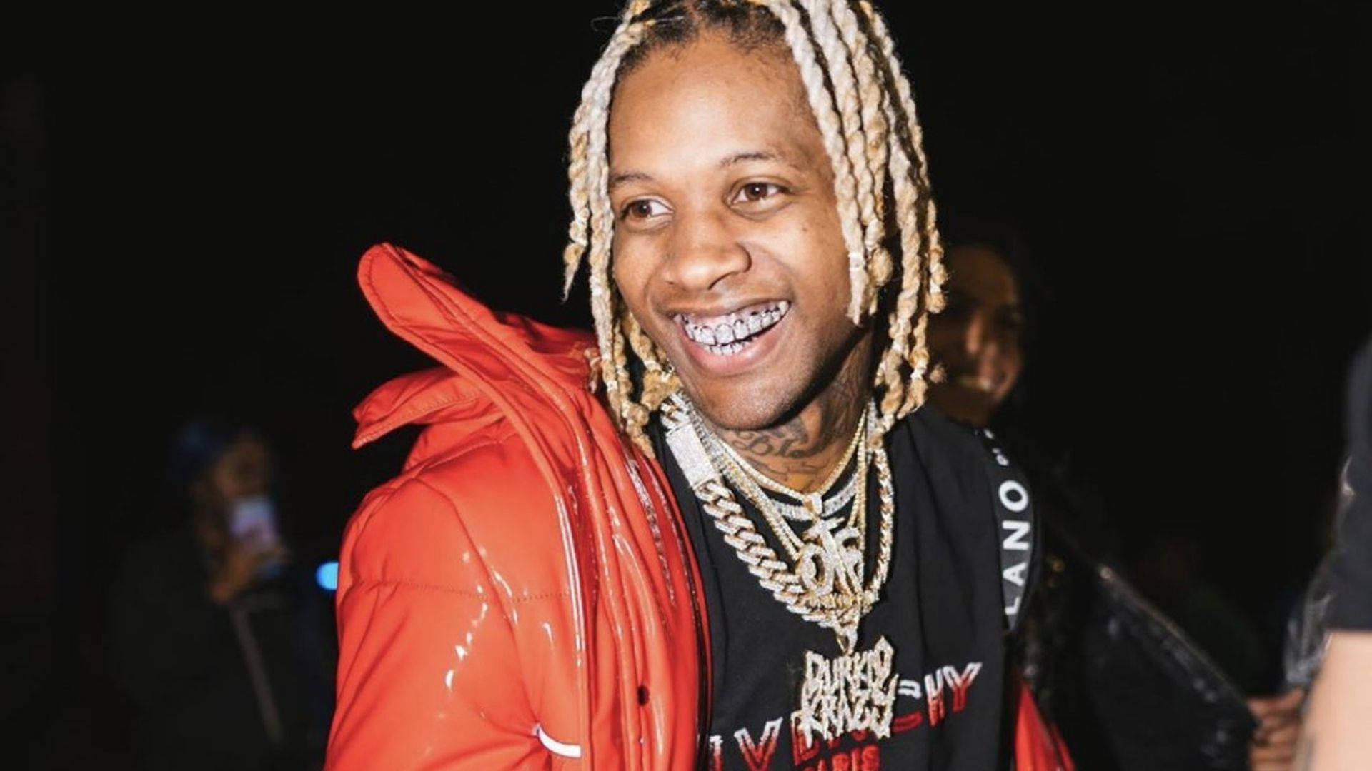 Lil Durk 桌布 壁紙 — 一位微笑的男子，編著辮子、戴著牙套，身穿亮紅色羽絨外套，層疊金鍊，背景為深色。