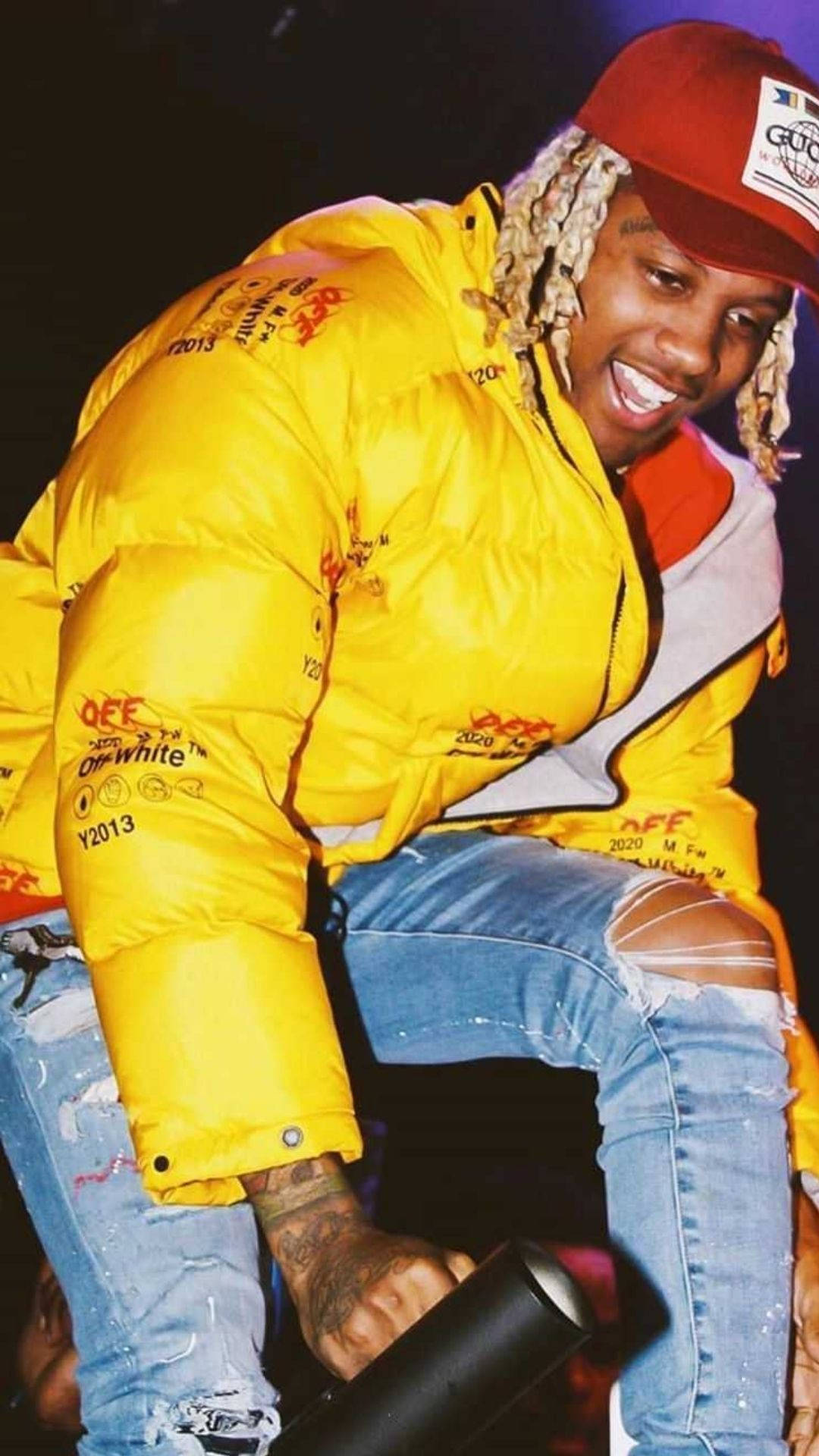 Lil Durk 桌布 壁紙 — 一位微笑的男子，編著辮子，身穿亮黃色羽絨外套和破洞牛仔褲，在舞台上拿著麥克風。