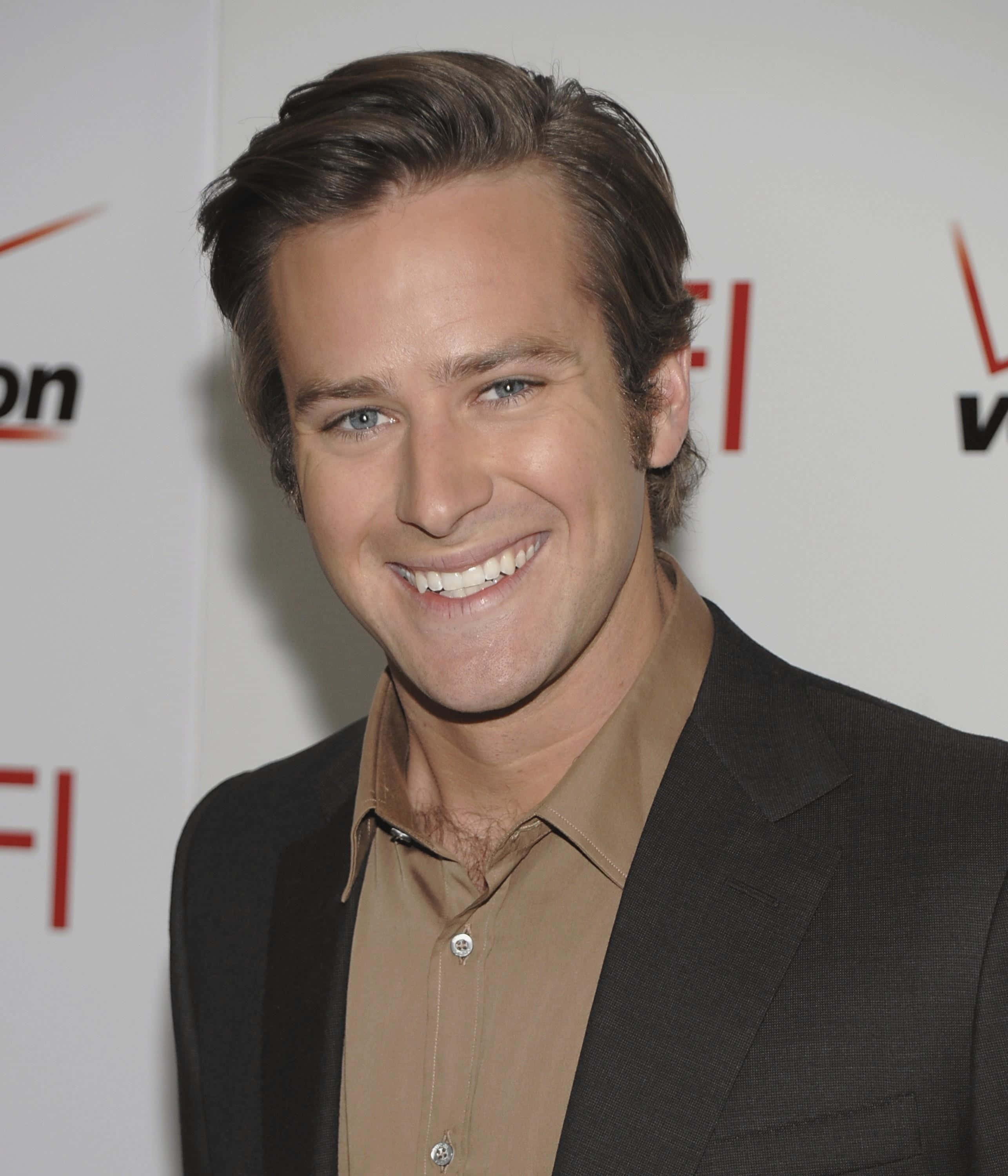 Armie Hammer 桌布 壁紙 — 一位微笑的男子，藍眼睛，棕色頭髮造型，身穿深色西裝和棕褐色襯衫，背景為品牌活動場景。