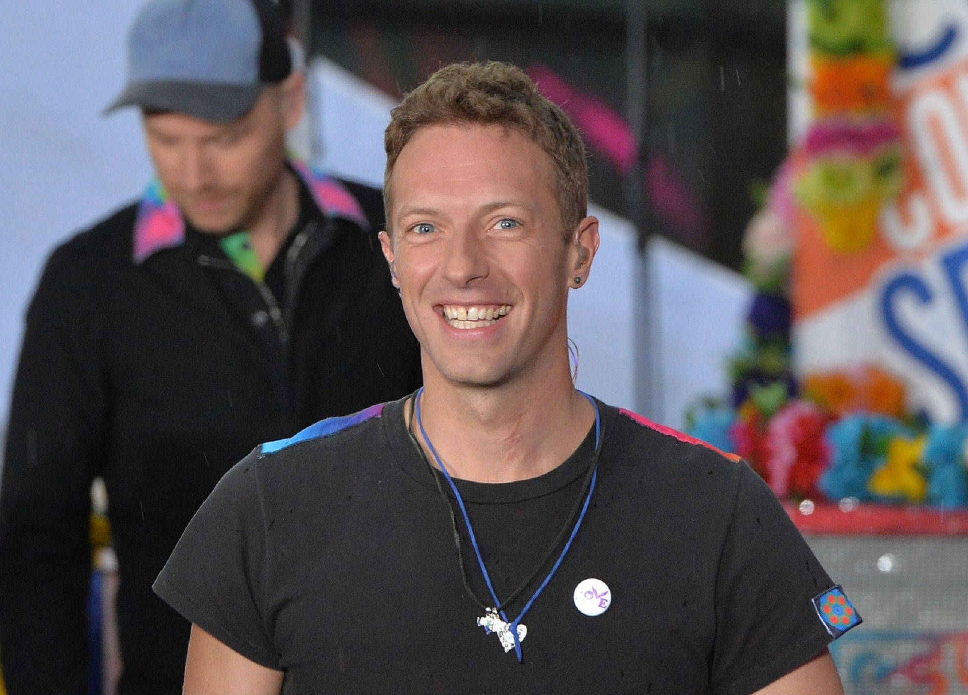 Chris Martin 桌布 壁紙 — 一位微笑的男子，藍眼睛，短棕髮，在色彩繽紛的活動中穿著黑色T恤和藍色掛繩。