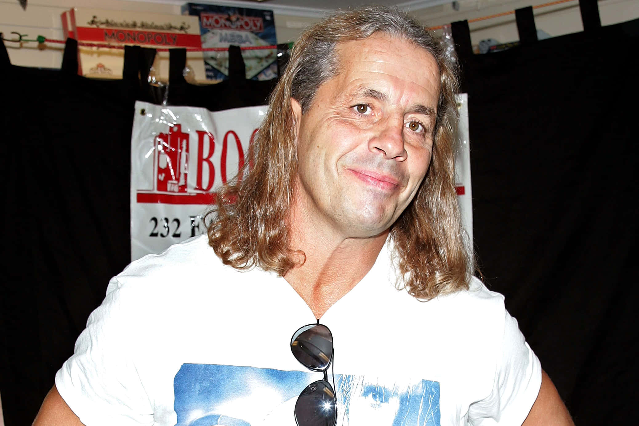 WWE 桌布 壁紙 — 一位微笑的男子，長捲髮，身穿白色T恤，衣領掛著太陽眼鏡，背景是活動橫幅。