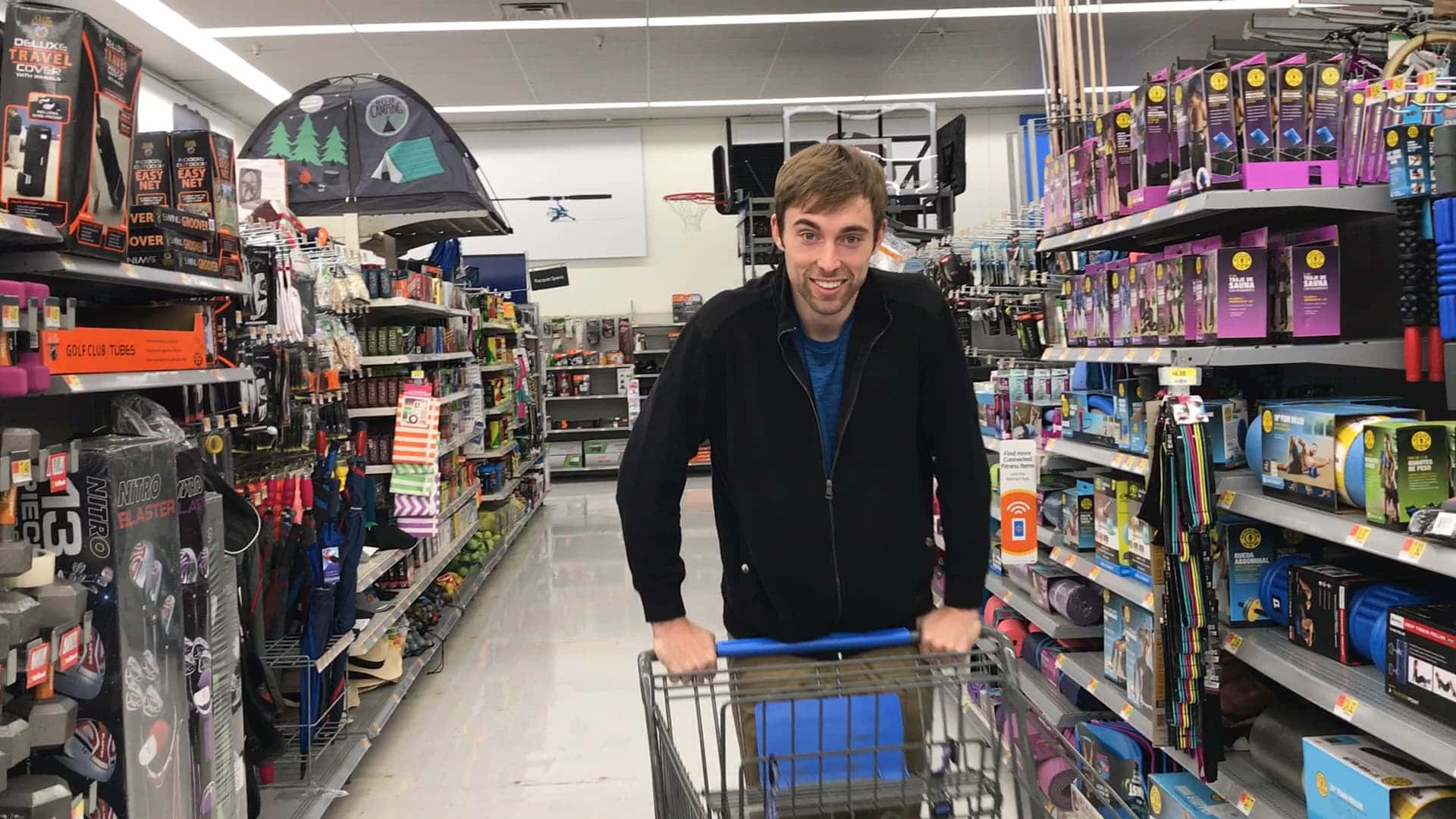 Shopping 桌布 壁紙 — 一位微笑的男子在充滿運動和露營裝備的零售店明亮走道中推著購物車。