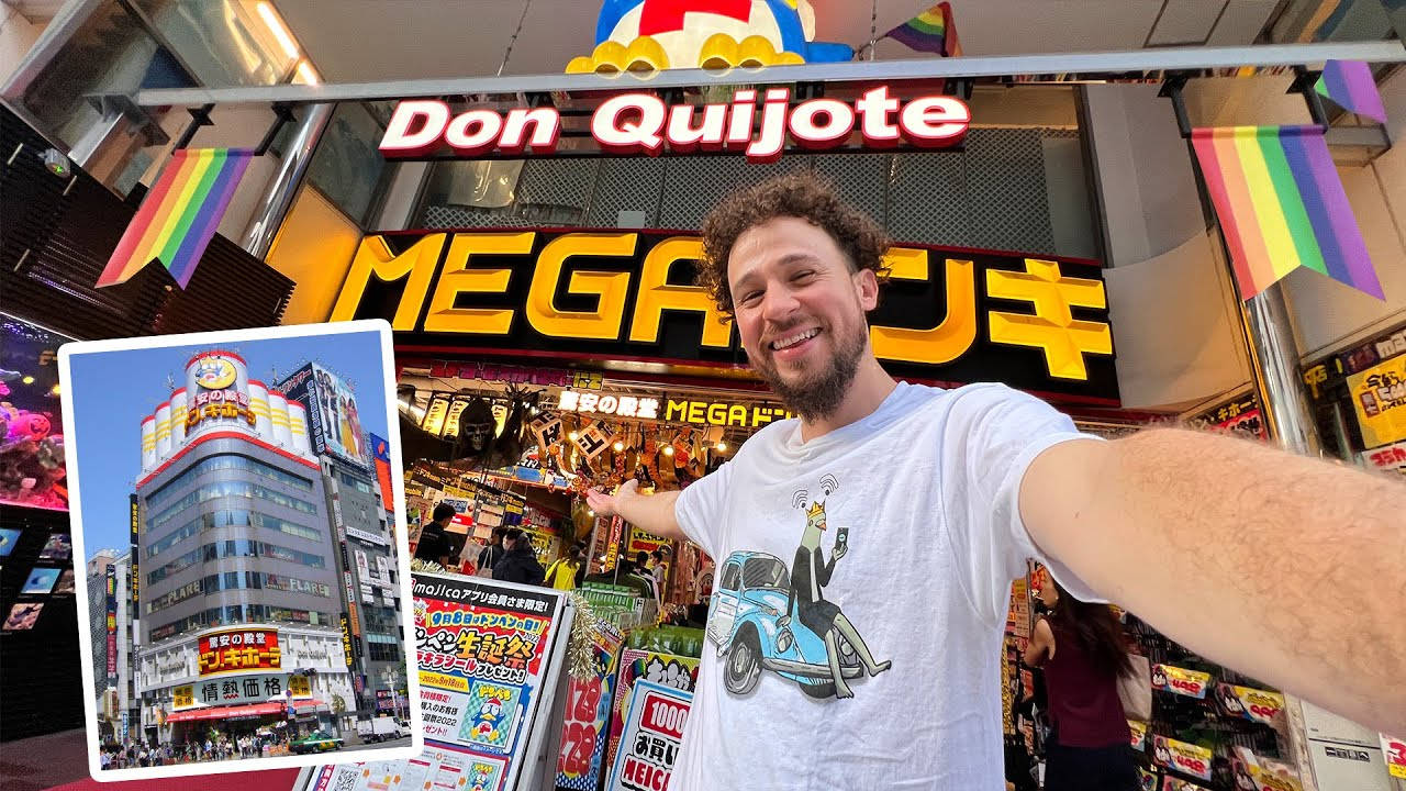 東京 桌布 壁紙 — 一位微笑的男子在燈光明亮的Don Quijote商店前自拍，附近展示著彩虹旗和建築物的照片。