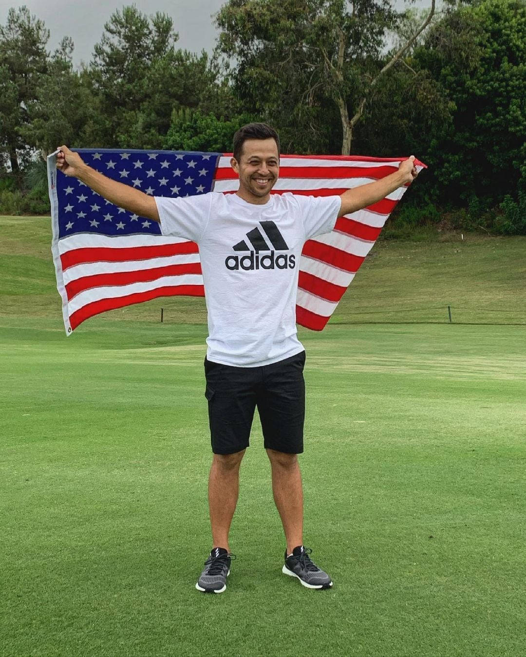 Flag 桌布 壁紙 — 一位微笑的男子在綠草如茵的高爾夫球場上自豪地揮舞著美國國旗，背景有樹木。