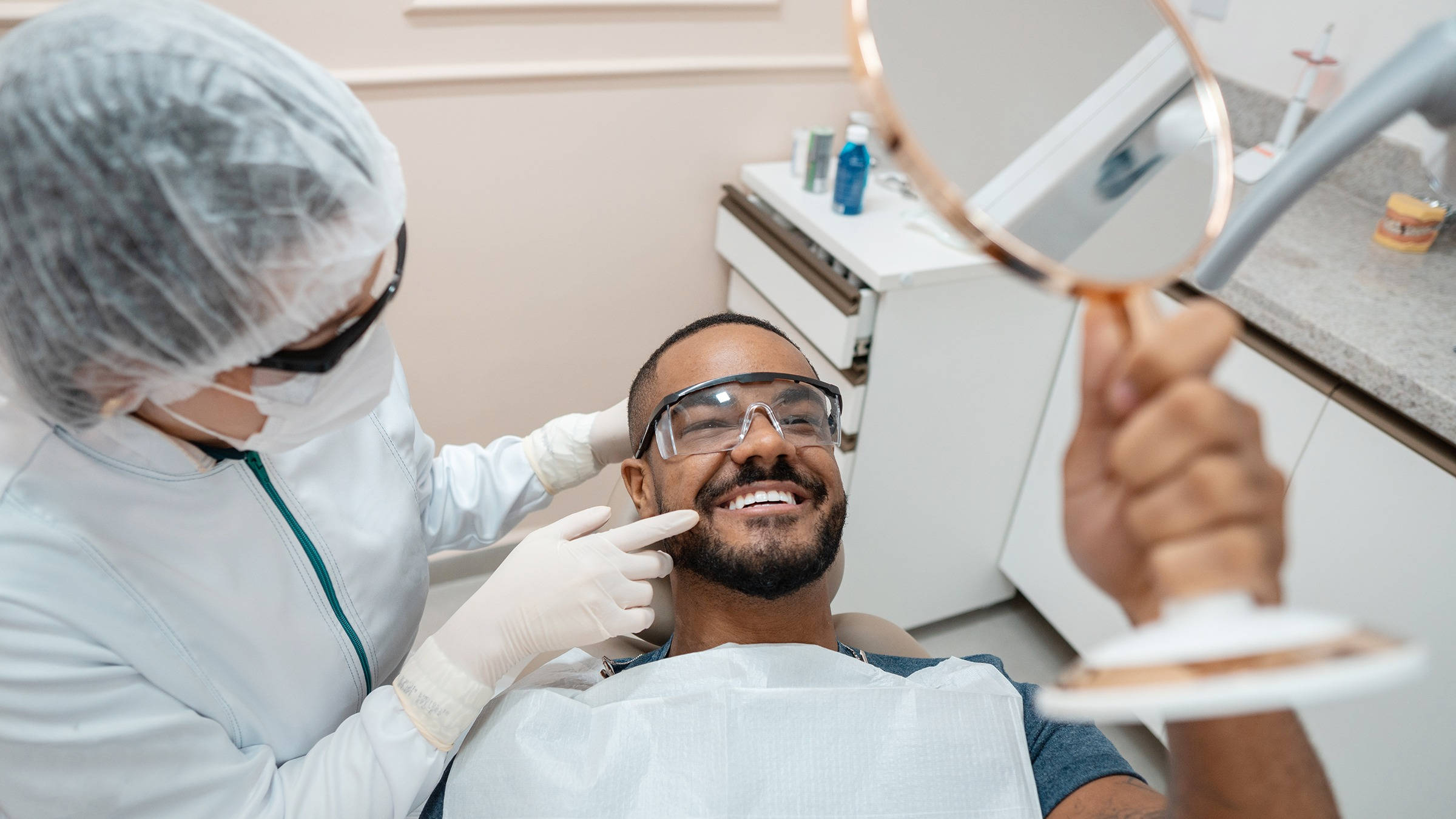 Dentistry 桌布 壁紙 — 一位微笑的男子坐在牙科椅上，用手鏡查看自己的新牙齒，而一位全副裝備的牙醫指著他的笑容。