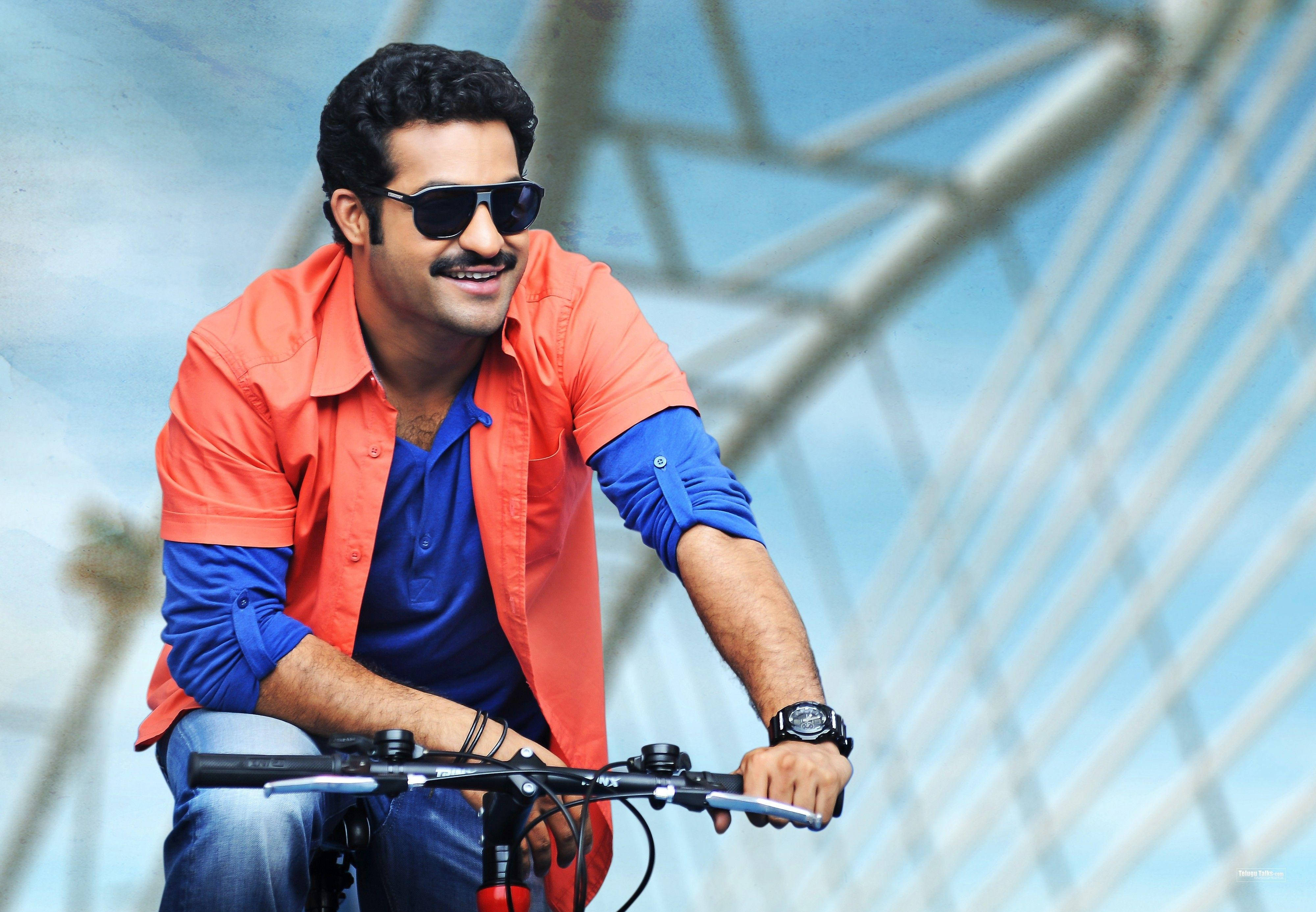 jr ntr 桌布 壁紙 — 一位微笑的男子戴著太陽眼鏡和彩色襯衫，在現代玻璃和鋼結構下騎自行車。