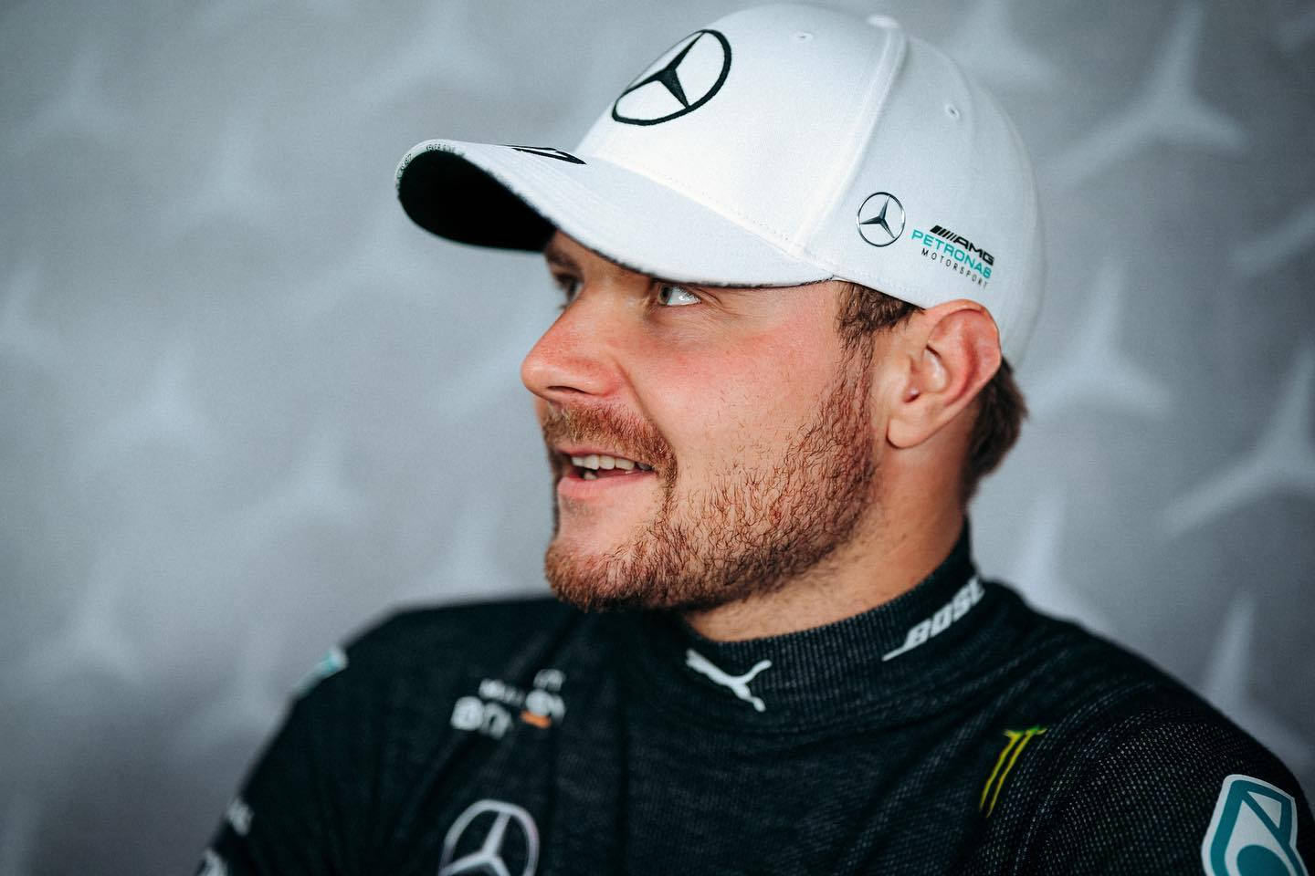 Valtteri Bottas 桌布 壁紙 — 一位微笑的男子戴著白色Mercedes帽子和黑色賽車服，在星形圖案的灰色背景前擺姿勢。