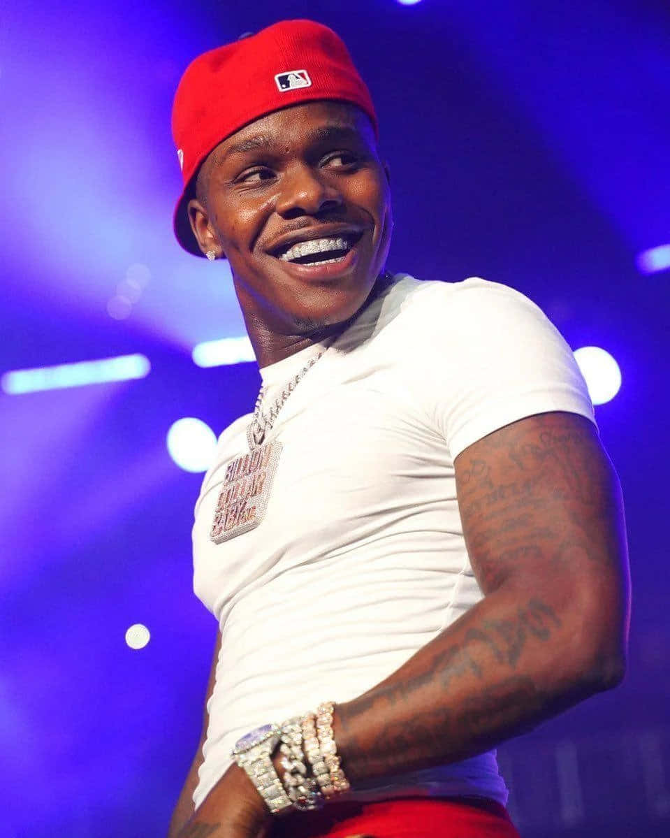 Dababy 桌布 壁紙 — 一位微笑的男子戴著紅色帽子、穿著白色襯衫，在充滿活力的藍色舞台燈光下表演，身上可見刺青和珠寶。