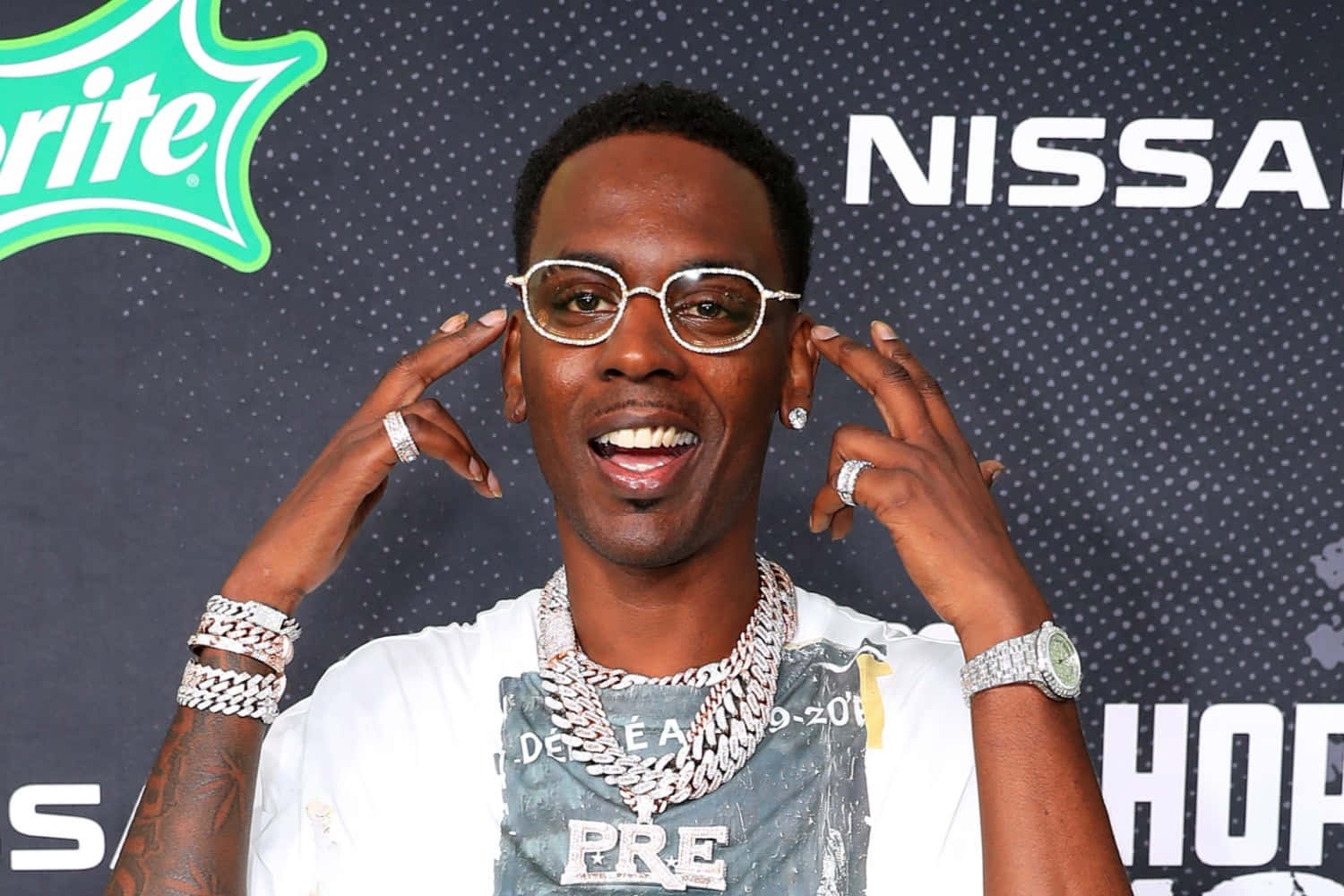 Young Dolph 桌布 壁紙 — 一位微笑的男子戴著閃亮珠寶和眼鏡，雙手靠近頭部，在品牌背景前擺姿勢。