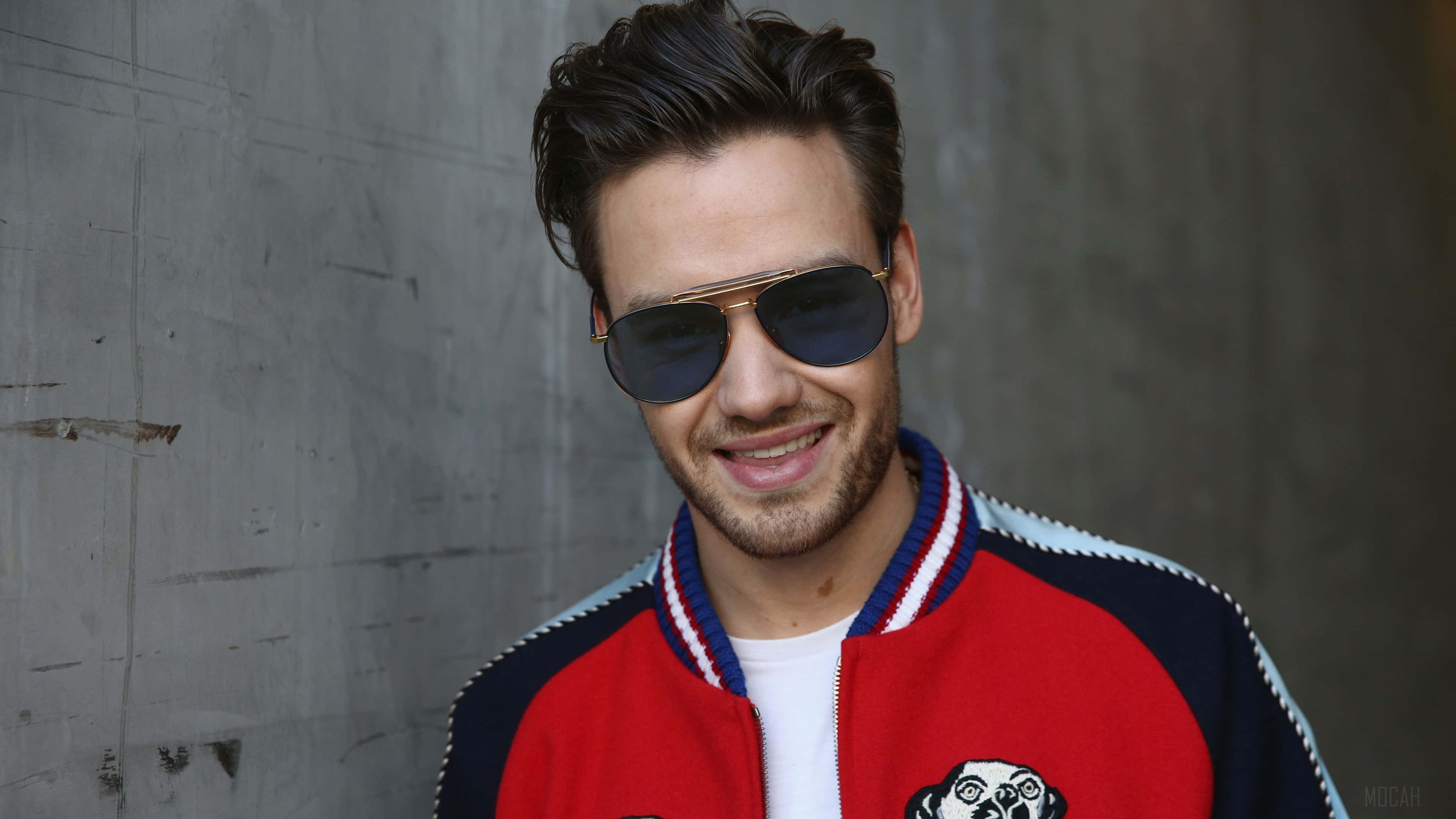 liam payne 桌布 壁紙 — 一位微笑的男子戴著飛行員太陽眼鏡，身穿紅藍色棒球外套，站在有紋理的灰色混凝土牆前。