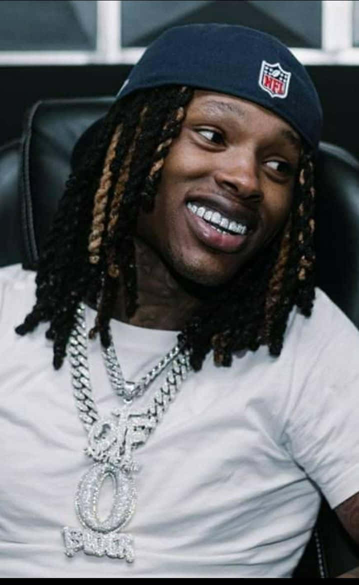 辮子男子戴NFL帽子和鑽石鏈條在黑色椅子上微笑。 — King Von 壁紙