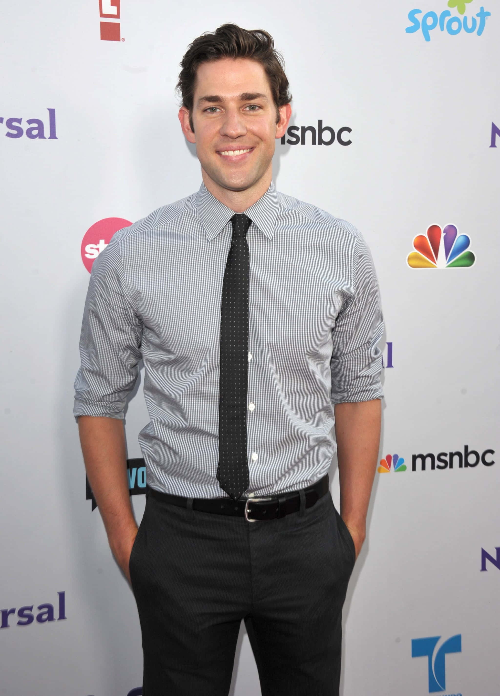 john krasinski 桌布 壁紙 — 一位微笑的男子穿著格子襯衫和黑色領帶，自信地站在媒體活動中，身後有標誌。