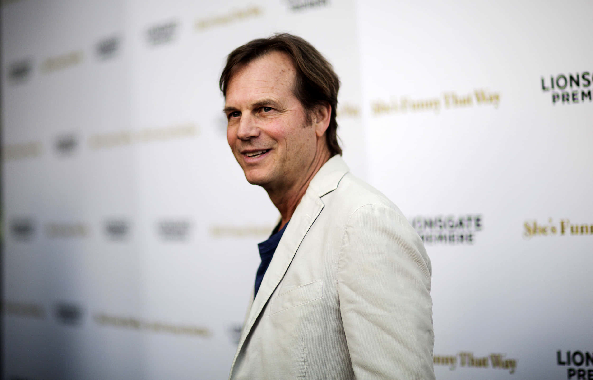 Celebrity 桌布 壁紙 — 一位微笑的男子穿著淺米色西裝外套，站在帶有標誌的品牌活動背景前。