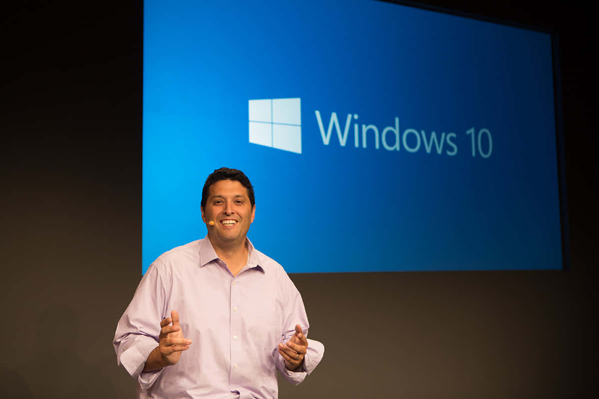 桌面 桌布 壁紙 — 一位微笑的男子穿著淺色襯衫，在顯示Windows 10標誌的大型藍色螢幕前做手勢進行簡報。