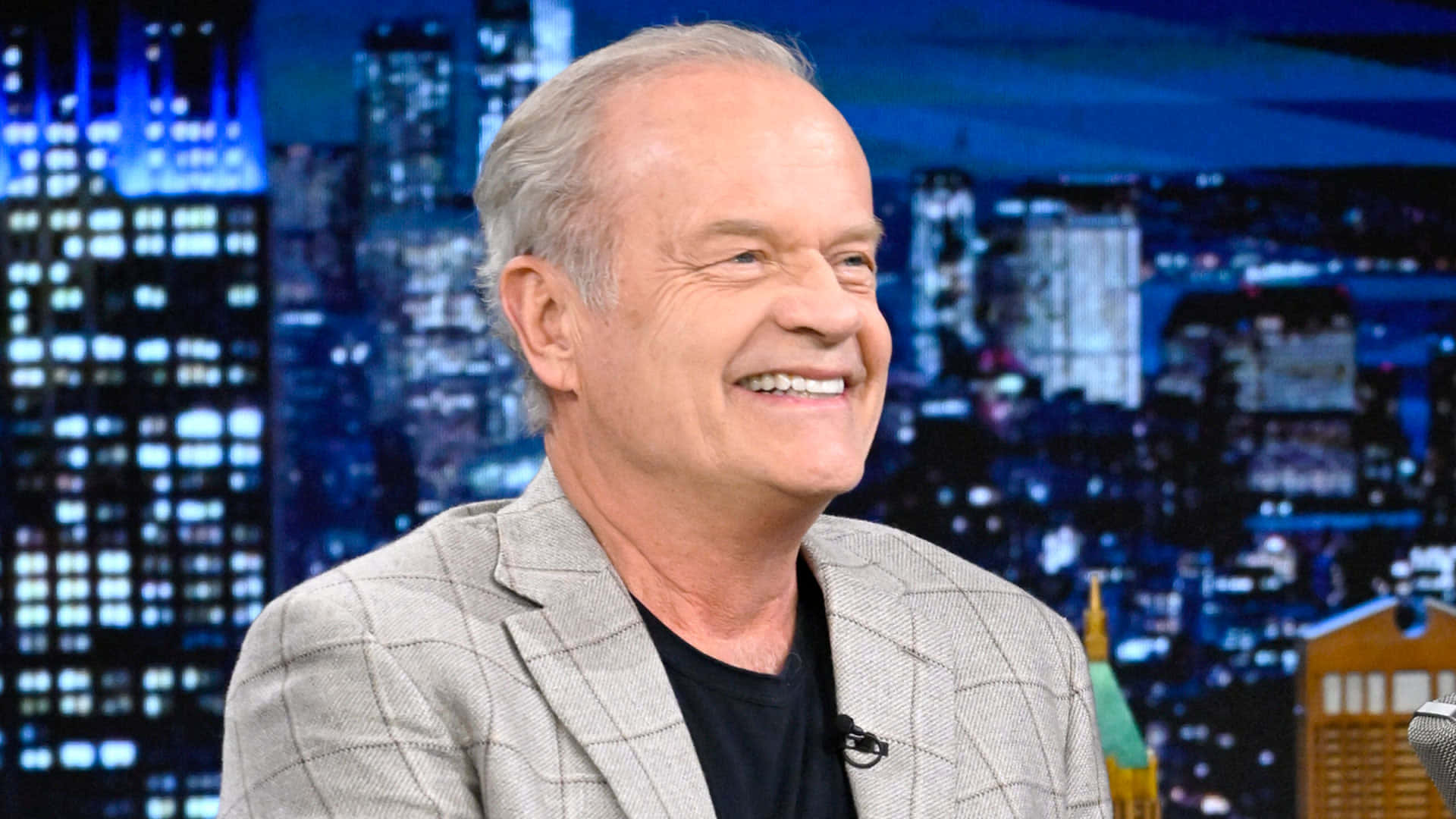 Kelsey Grammer 桌布 壁紙 — 一位微笑的男子穿著灰色西裝外套，在充滿活力的藍色夜景城市背景下溫暖地笑著。