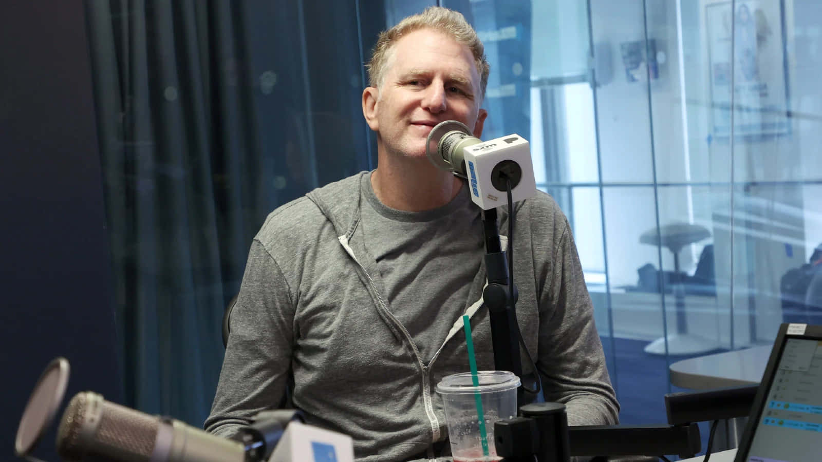Michael Rapaport 桌布 壁紙 — 一位微笑的男子穿著灰色連帽衫，坐在現代玻璃牆工作室的桌子旁，桌上有麥克風和一個Starbucks杯子。