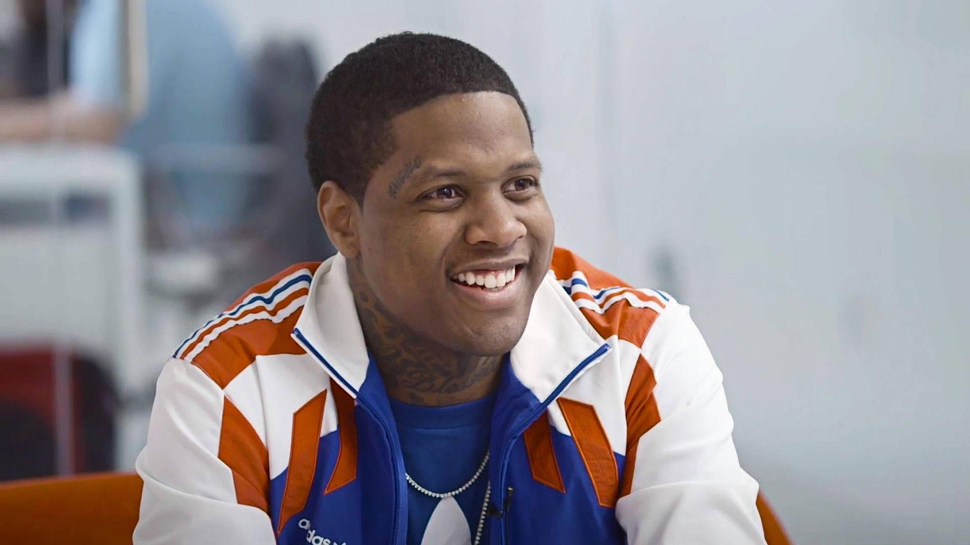 Lil Durk 桌布 壁紙 — 一位微笑的男子穿著白色、橙色和藍色的Adidas運動外套，露出頸部刺青，坐在室內。