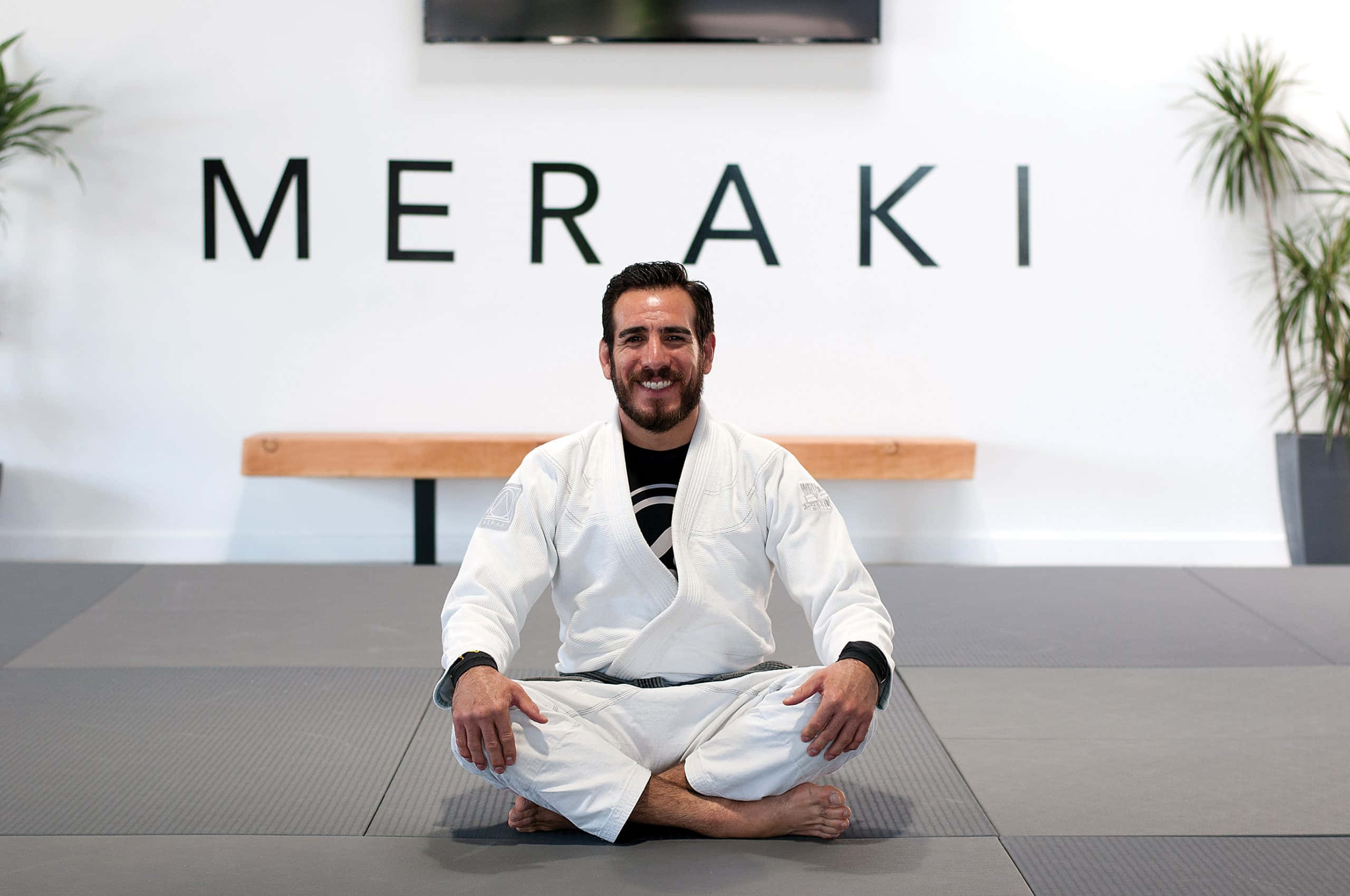 Jiu-jitsu 桌布 壁紙 — 一位微笑的男子穿著白色柔道服，在明亮的現代工作室中盤腿坐在灰色墊子上，牆上寫著「MERAKI」。