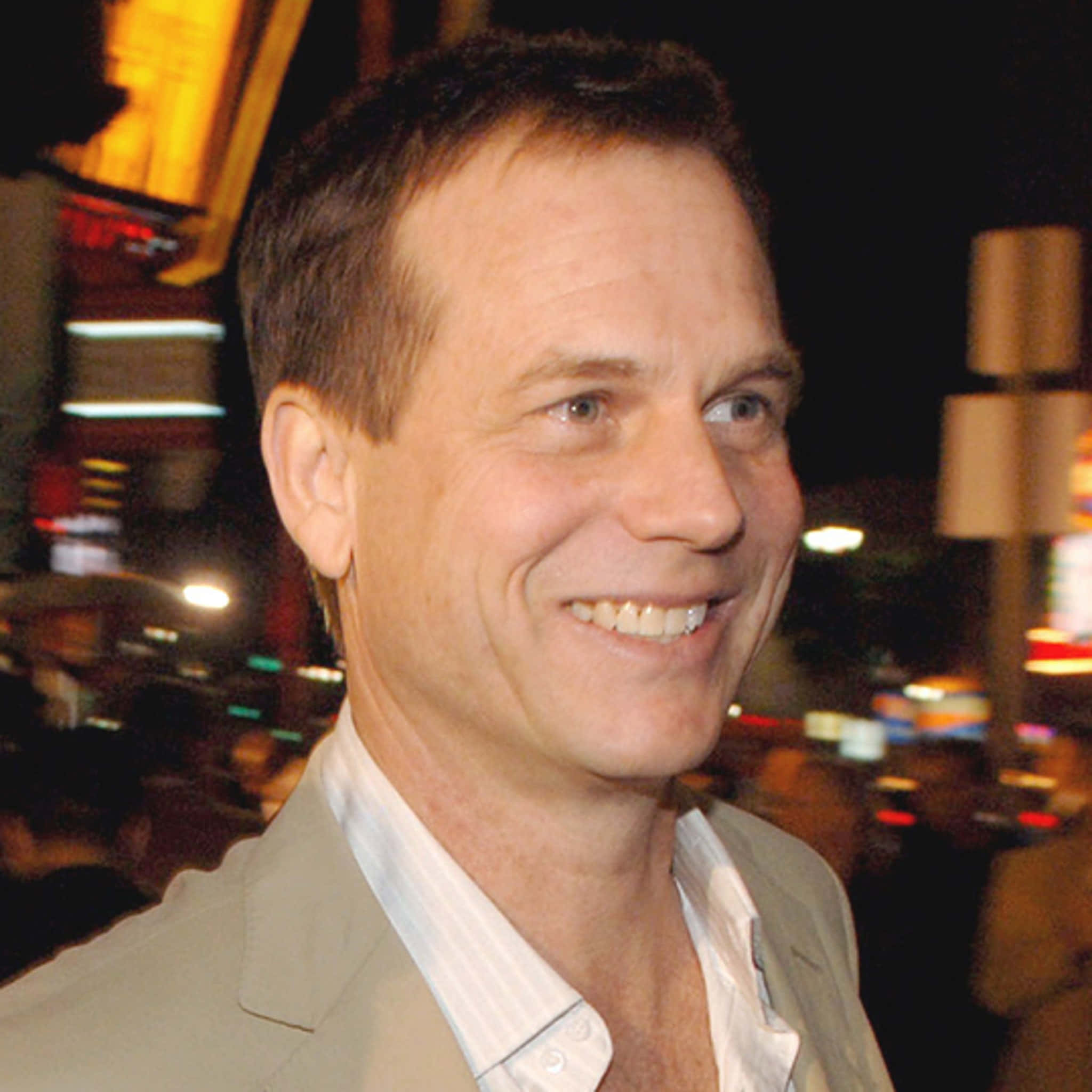 actor 桌布 壁紙 — 一位微笑的男子穿著米色西裝外套和白色襯衫，站在模糊的夜間城市景觀前，背景有閃爍的燈光。