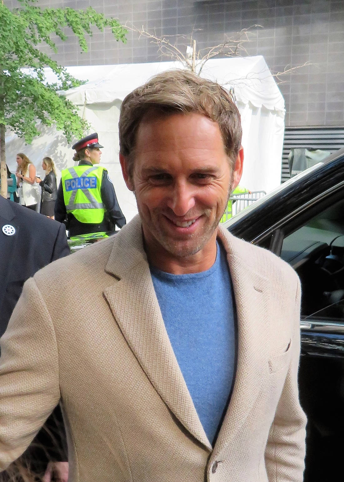 Celebrity 桌布 壁紙 — 一位微笑的男子穿著米色西裝外套和藍色襯衫，站在戶外，附近有一名警察和一輛黑色汽車。