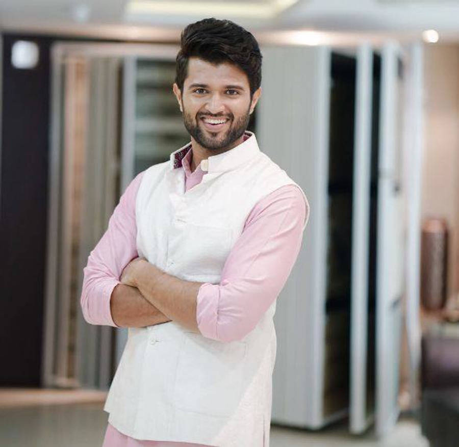 vijay devarakonda 4K 桌布 壁紙 — 一位微笑的男子穿著粉紅色襯衫和白色背心，自信地交叉雙臂站在現代室內空間中。