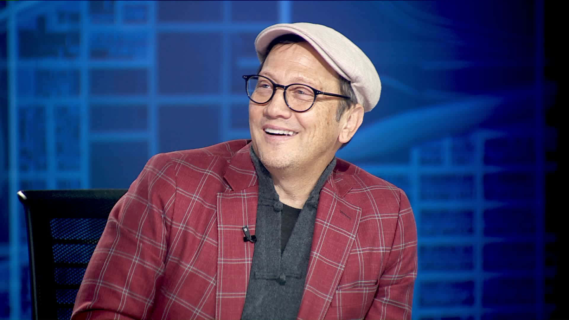 actor 桌布 壁紙 — 一位微笑的男子穿著紅色格紋西裝外套和米色帽子，在藍色網格圖案的工作室背景前溫暖地笑著。