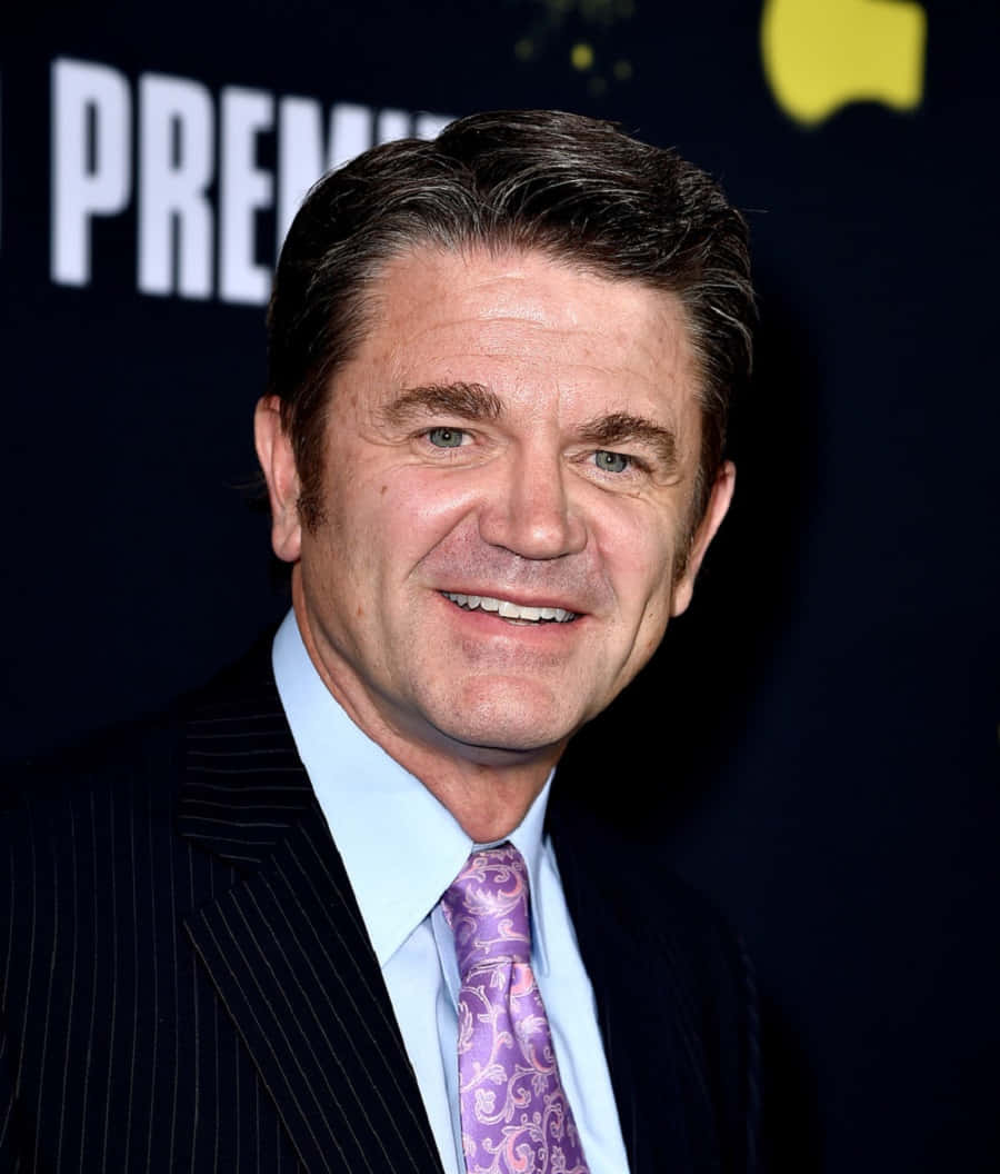 John Michael Higgins 桌布 壁紙 — 一位微笑的男子穿著細條紋西裝和圖案紫色領帶，在帶有 'PREMIERE' 文字的深色背景前擺姿勢。