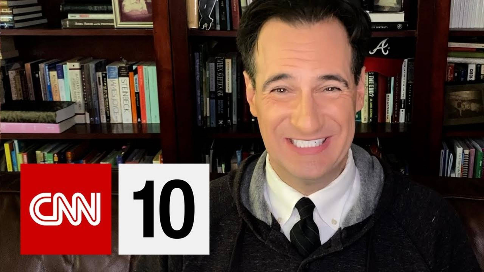 tv 桌布 壁紙 — 一位微笑的男子穿著連帽衫和領帶，坐在一個顯示CNN 10標誌的書架前。