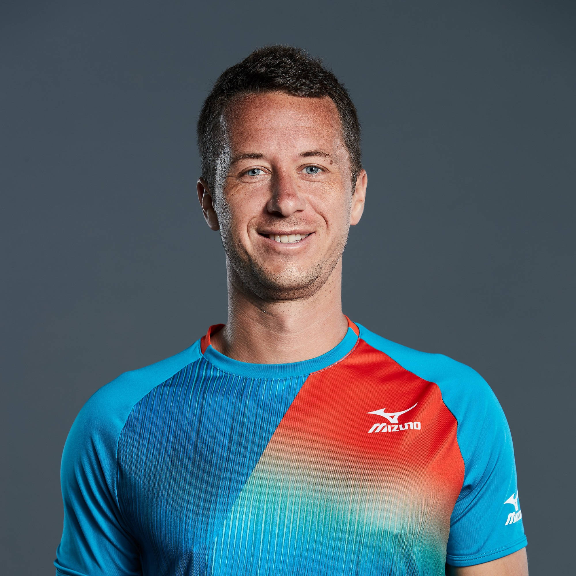 人像動態 桌布 壁紙 — 一位微笑的男子穿著鮮豔的藍紅 Mizuno 運動衫，在純灰色工作室背景前擺姿勢。