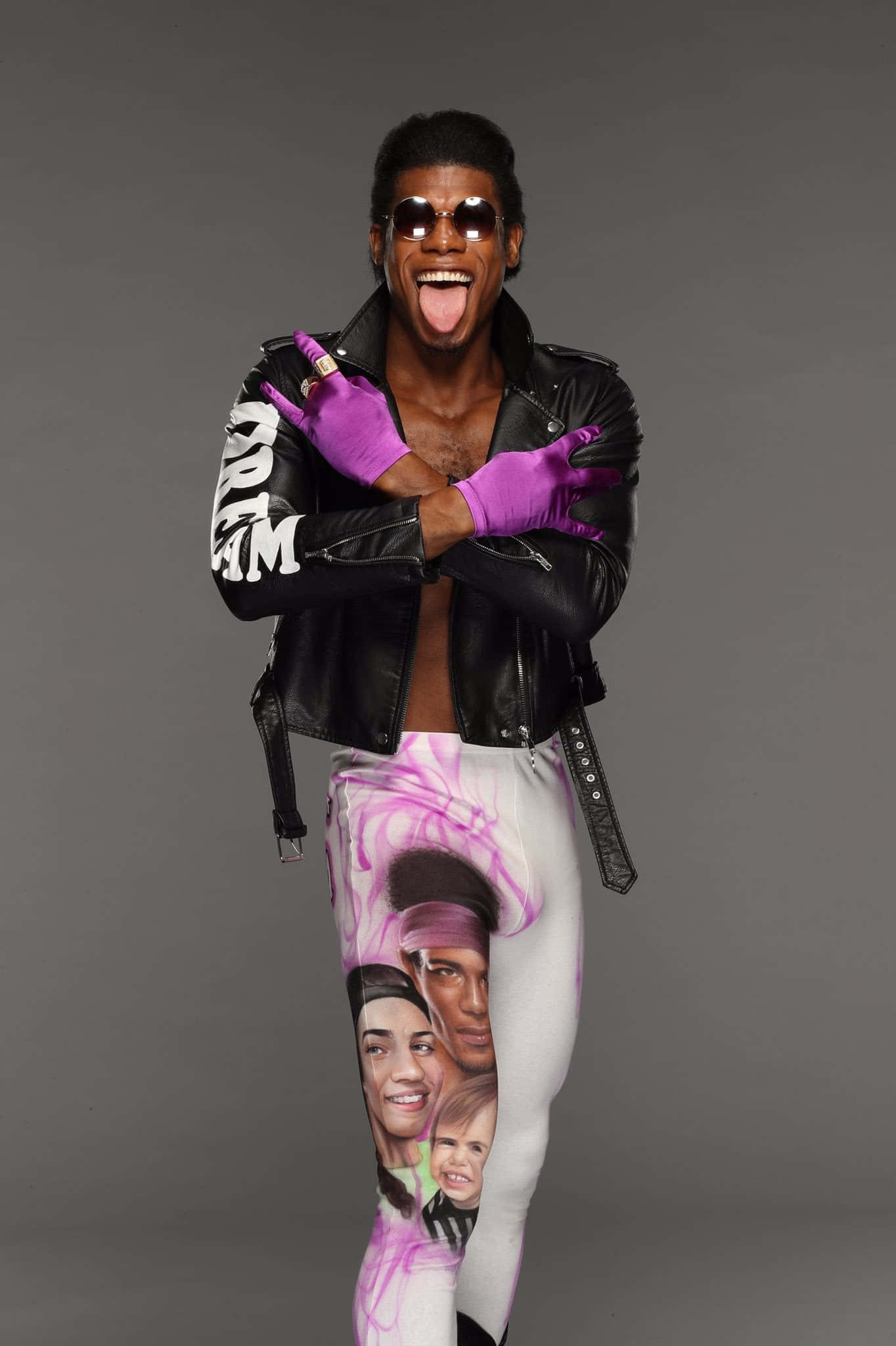 WWE 桌布 壁紙 — 一位微笑的男子穿著黑色皮夾克和紫色手套，穿著印有臉孔的緊身褲，在灰色攝影棚背景下伸出舌頭。