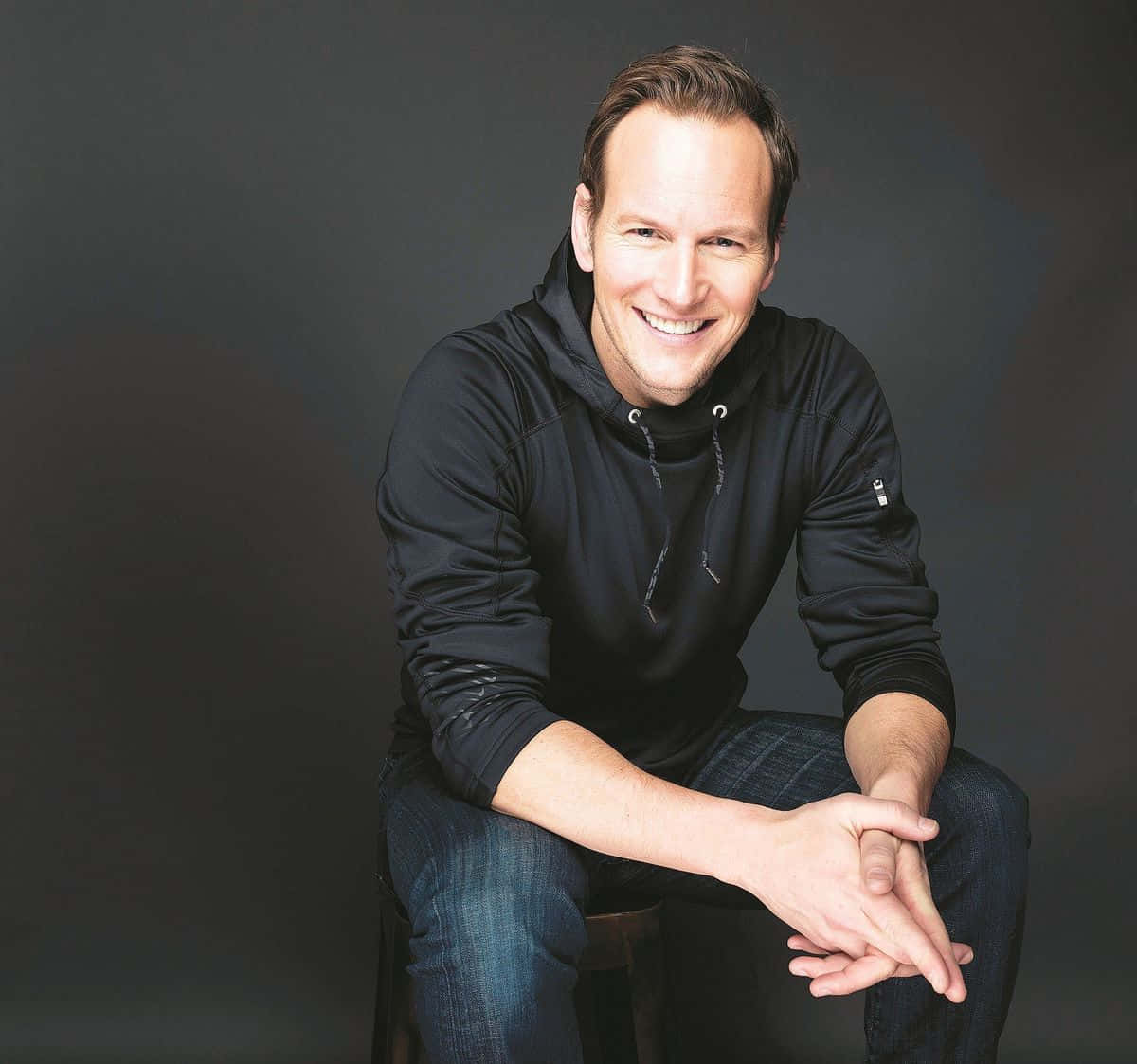 Patrick Wilson 桌布 壁紙 — 一位微笑的男子穿著黑色連帽衫和牛仔褲，隨意地坐在深灰色攝影棚背景前的凳子上。