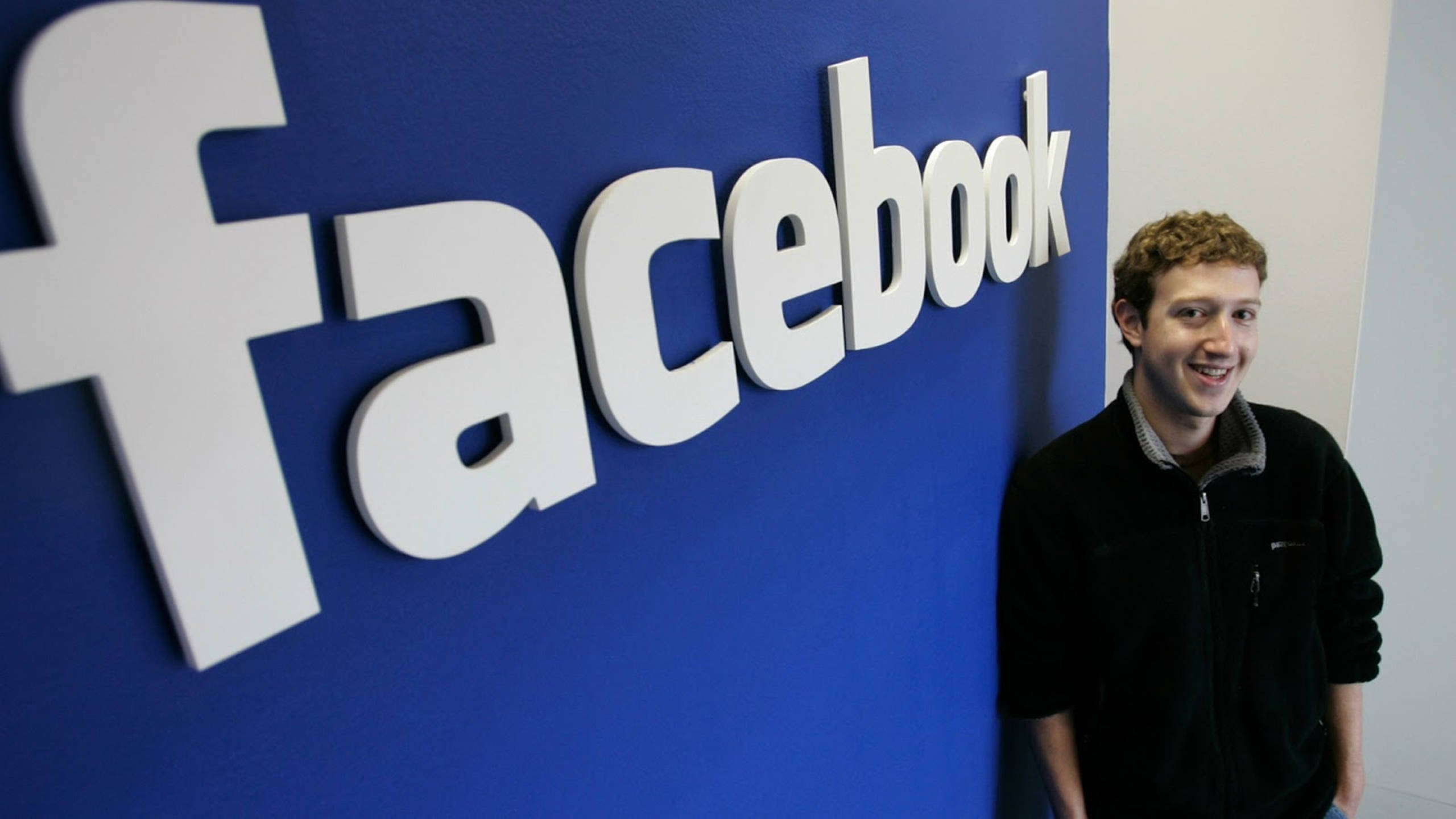 Zuckerberg 桌布 壁紙 — 一位微笑的男子站在辦公室環境中，旁邊是一個安裝在鮮豔藍色牆上的大型白色Facebook標誌。