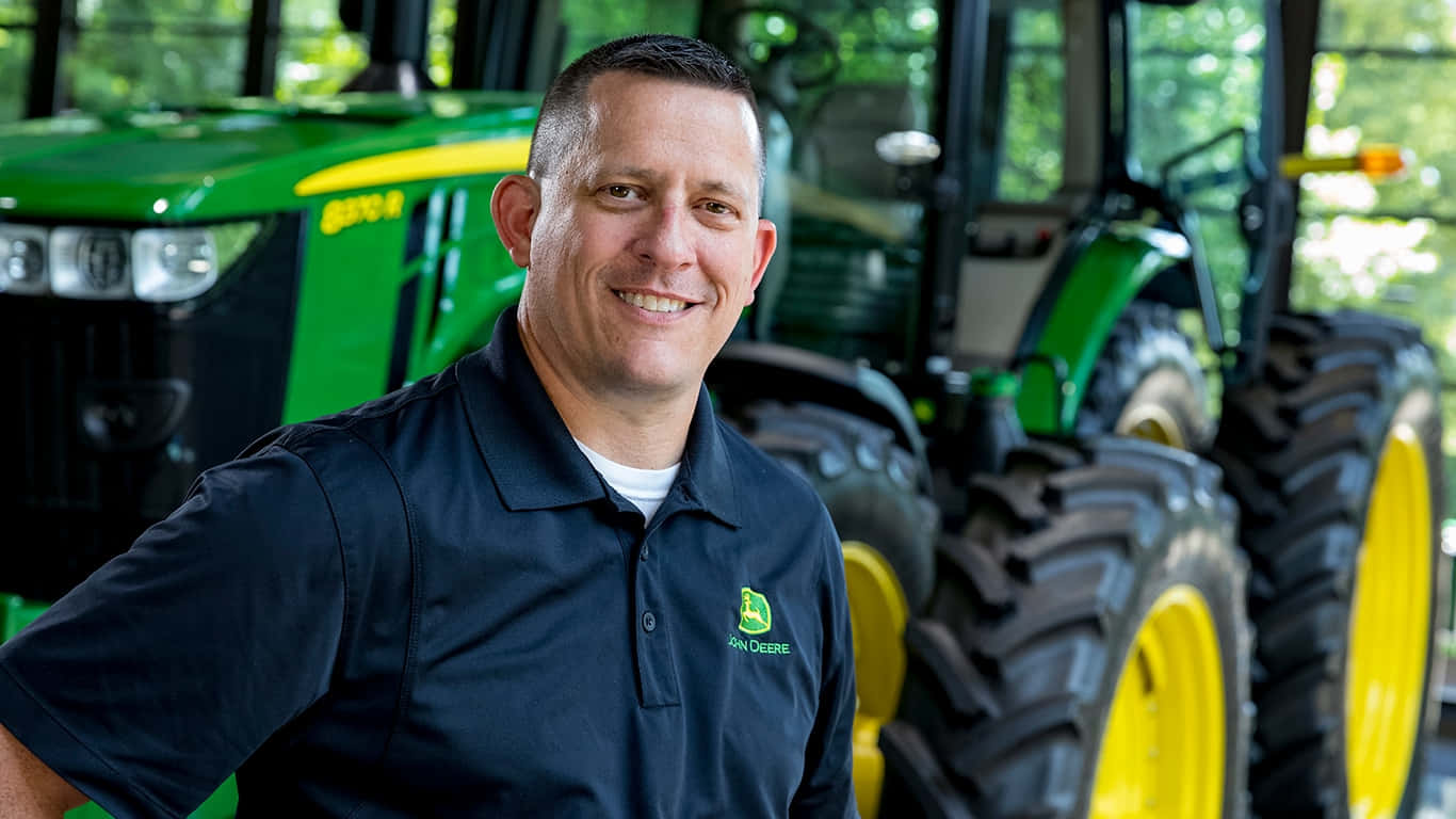 約翰迪爾 桌布 壁紙 — 一位微笑的男子身穿John Deere Polo衫，自信地站在一輛帶有大型黑色輪胎的綠色和黃色拖拉機前。