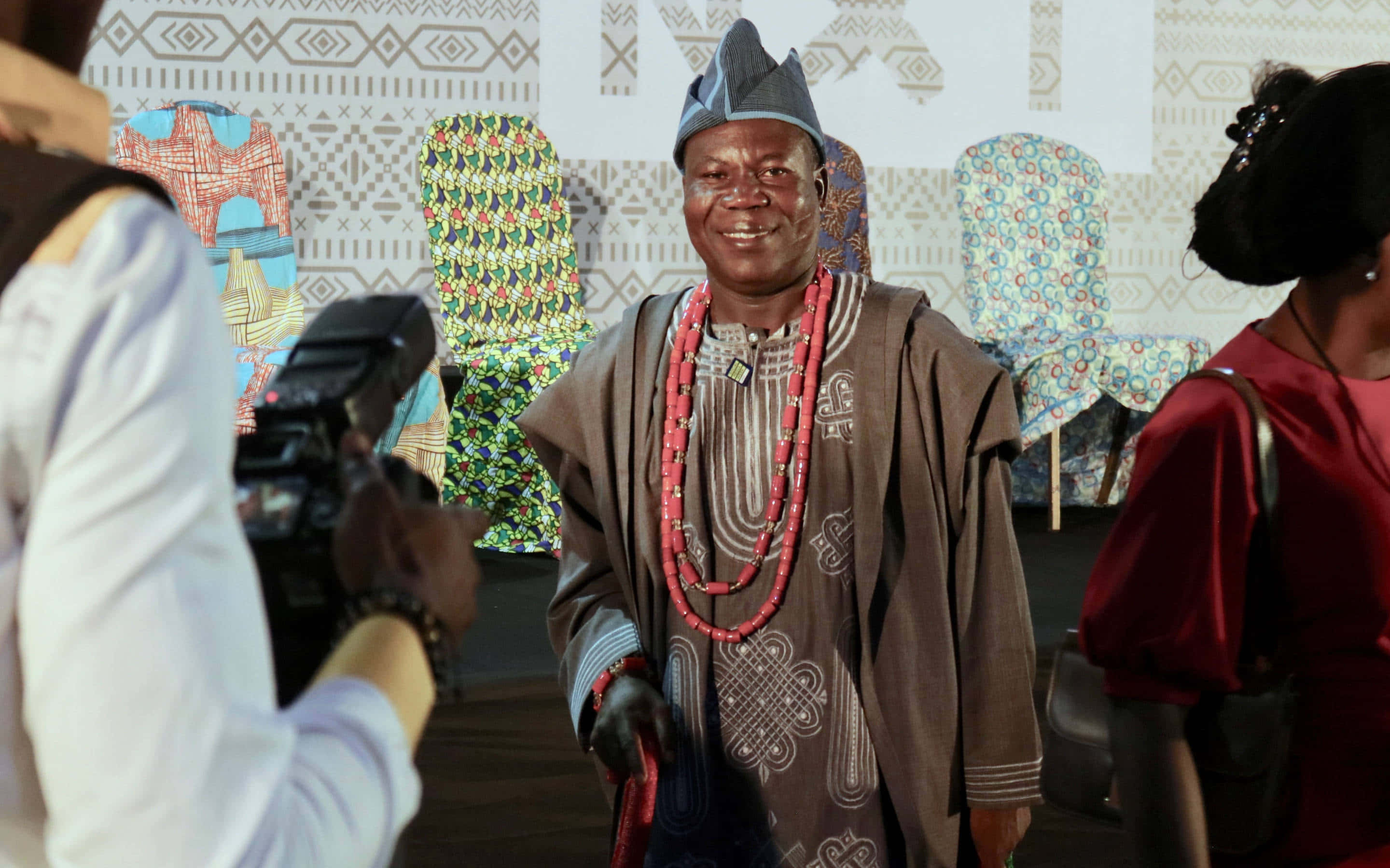 Nigeria 桌布 壁紙 — 一位微笑的男子身穿傳統奈及利亞服飾，配戴紅色珠子和圖案布料，站在文化活動中，攝影師捕捉他的瞬間。