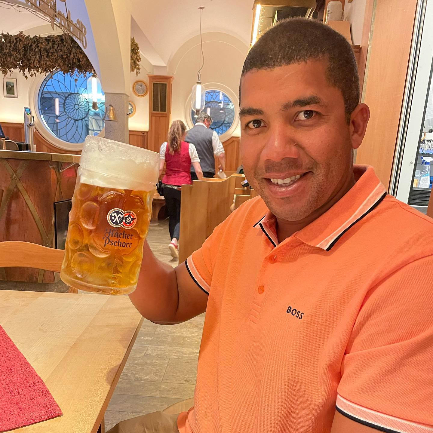 Beer 桌布 壁紙 — 一位微笑的男子身穿橙色Polo衫，在舒適的木製酒吧自豪地舉起一大杯泡沫豐富的Hacker Pschorr啤酒杯。