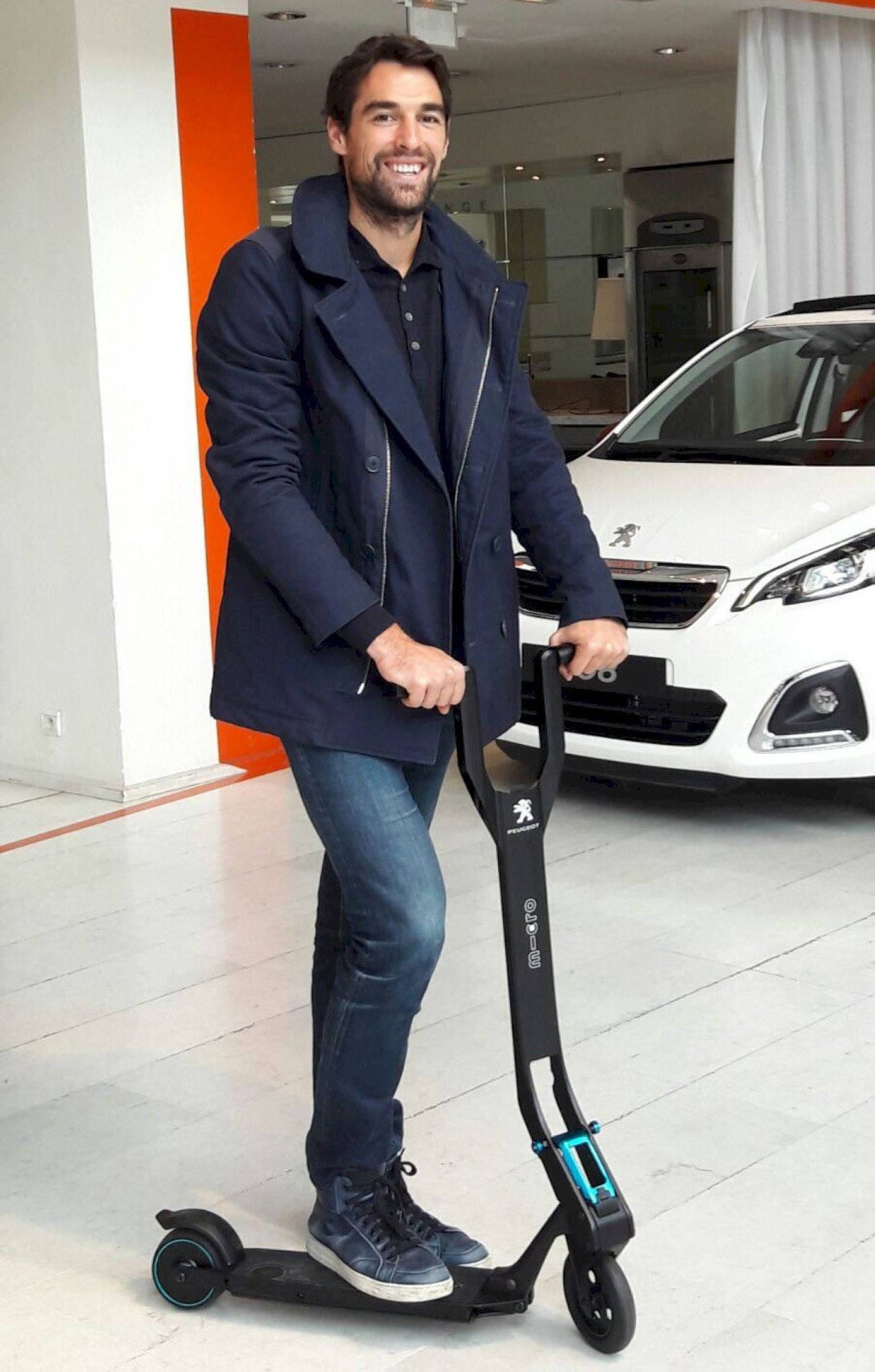 scooter 桌布 壁紙 — 一位微笑的男子身穿海軍藍外套，自信地站在黑色電動滑板車上，身處現代汽車經銷店內，身後有一輛白色標緻汽車。
