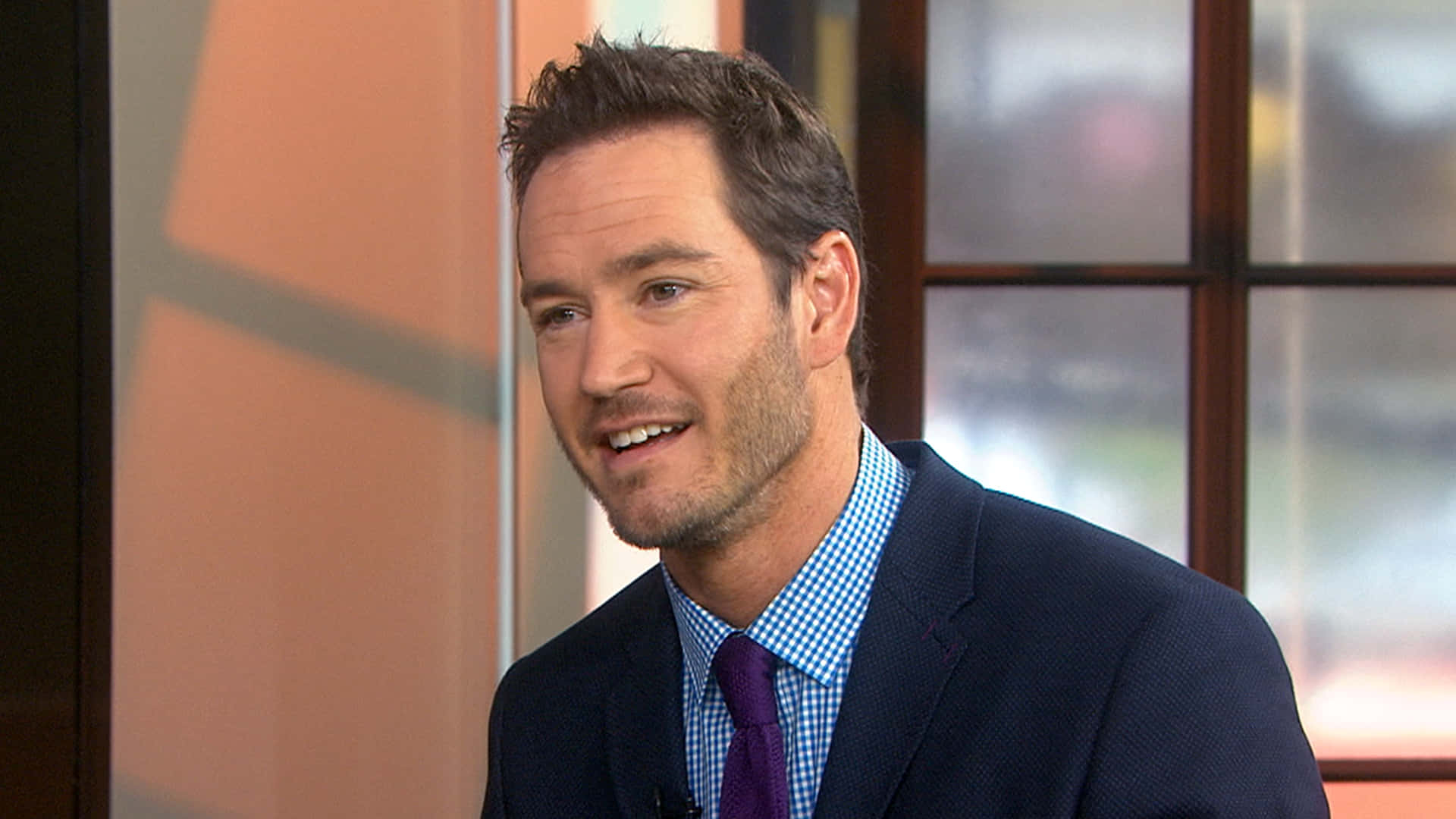 Mark-paul Gosselaar 桌布 壁紙 — 一位微笑的男子身穿海軍藍西裝和紫色領帶，在白天的工作室訪談中說話，身後有柔和的窗光。