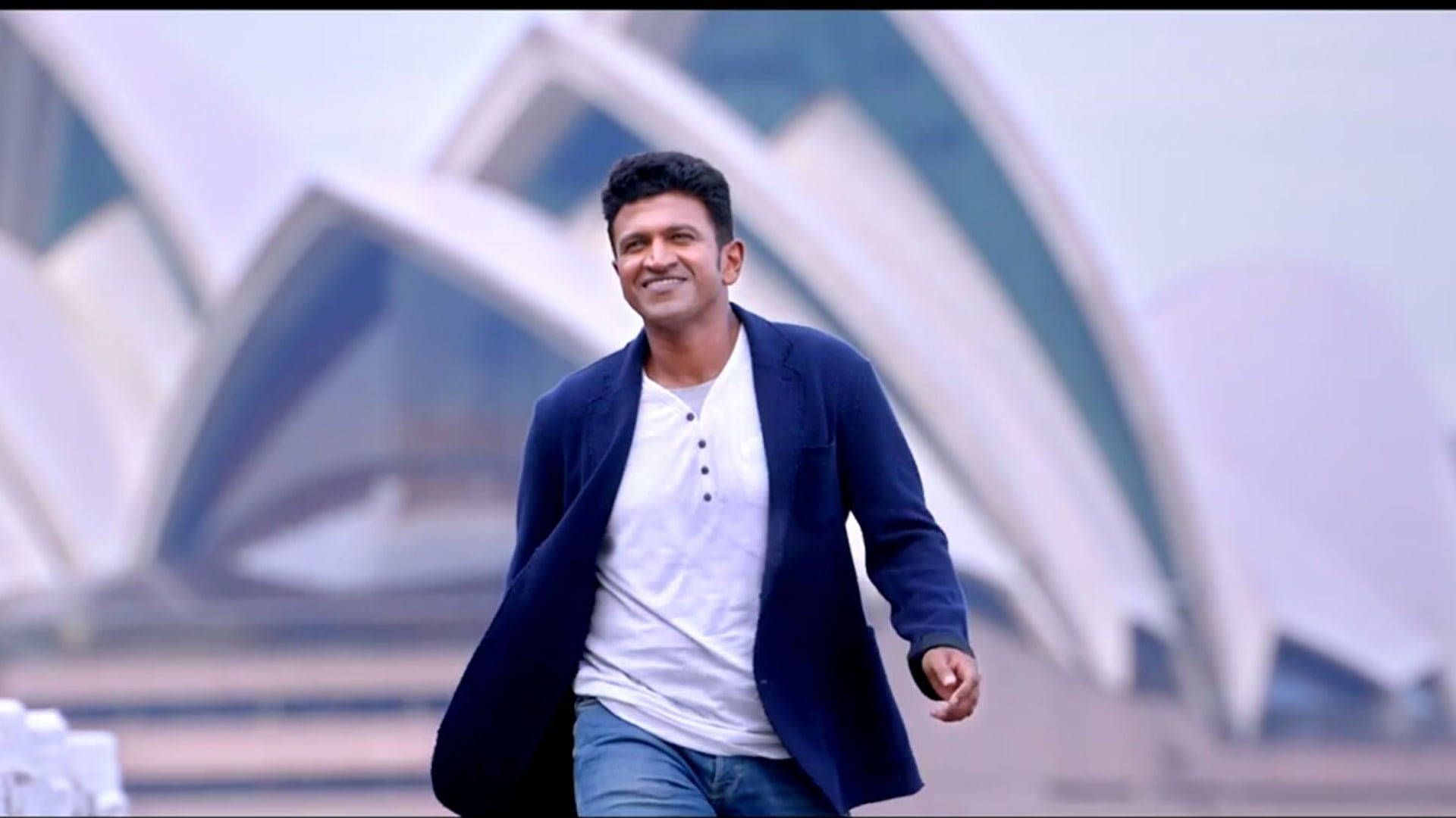 Puneeth Rajkumar 桌布 壁紙 — 一位微笑的男子身穿海軍藍西裝外套和牛仔褲，自信地走在標誌性的雪梨歌劇院前。