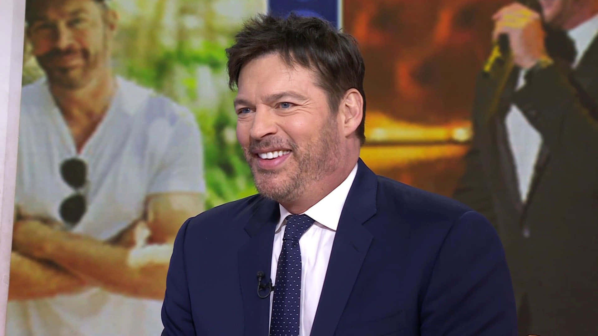 Harry Connick Jr 桌布 壁紙 — 一位微笑的男子身穿海軍藍西裝搭配波點領帶，在模糊的宣傳背景佈景中發言。