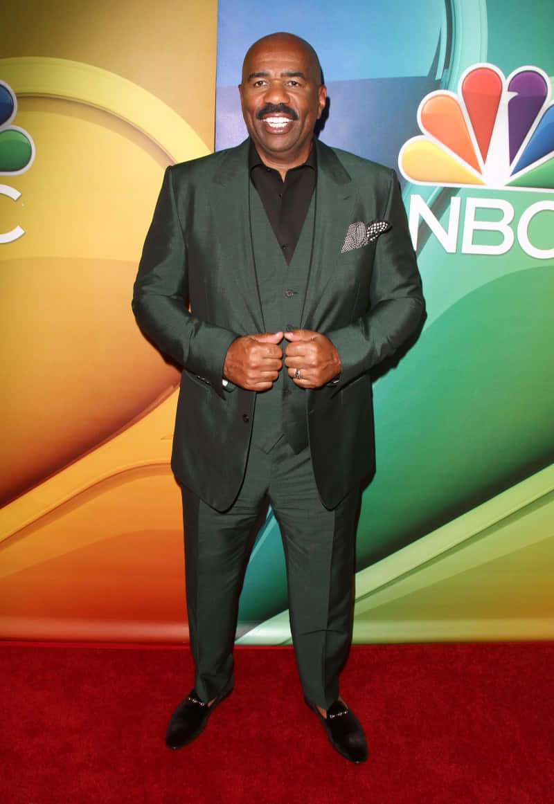 steve harvey 桌布 壁紙 — 一位微笑的男子身穿深綠色西裝，自信地站在紅地毯上，背景是色彩繽紛的NBC標誌。