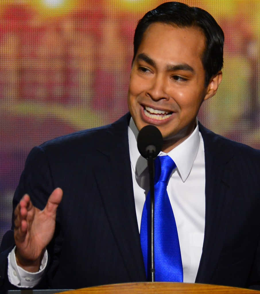 Celebrity 桌布 壁紙 — 一位微笑的男子身穿深色西裝和亮藍色領帶，在舞台上對著麥克風說話並做手勢，身後有溫暖模糊的燈光。