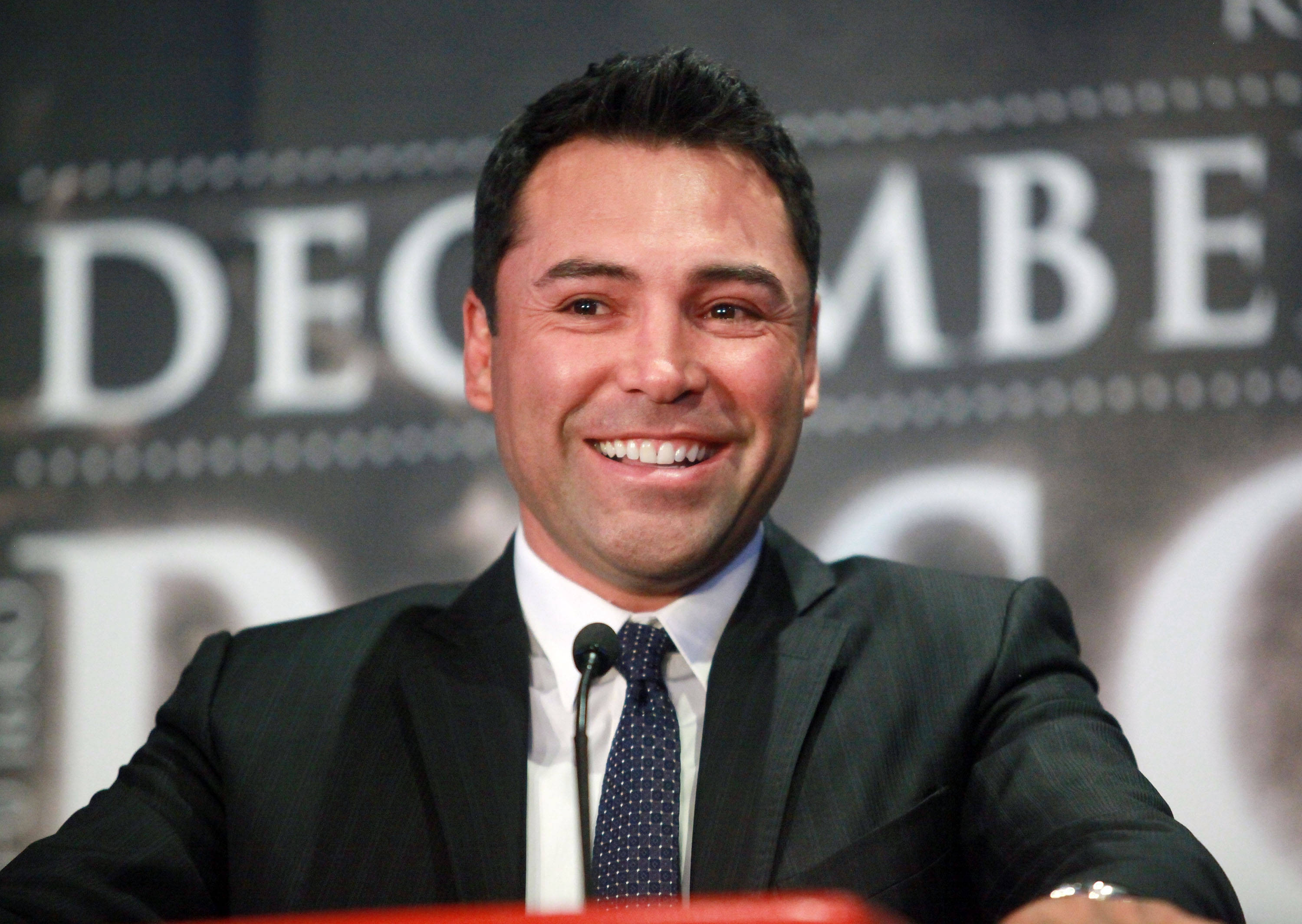 Boxing 桌布 壁紙 — 一位微笑的男子身穿深色西裝和圖案領帶，坐在有'DECEMBER'背景的講台上。