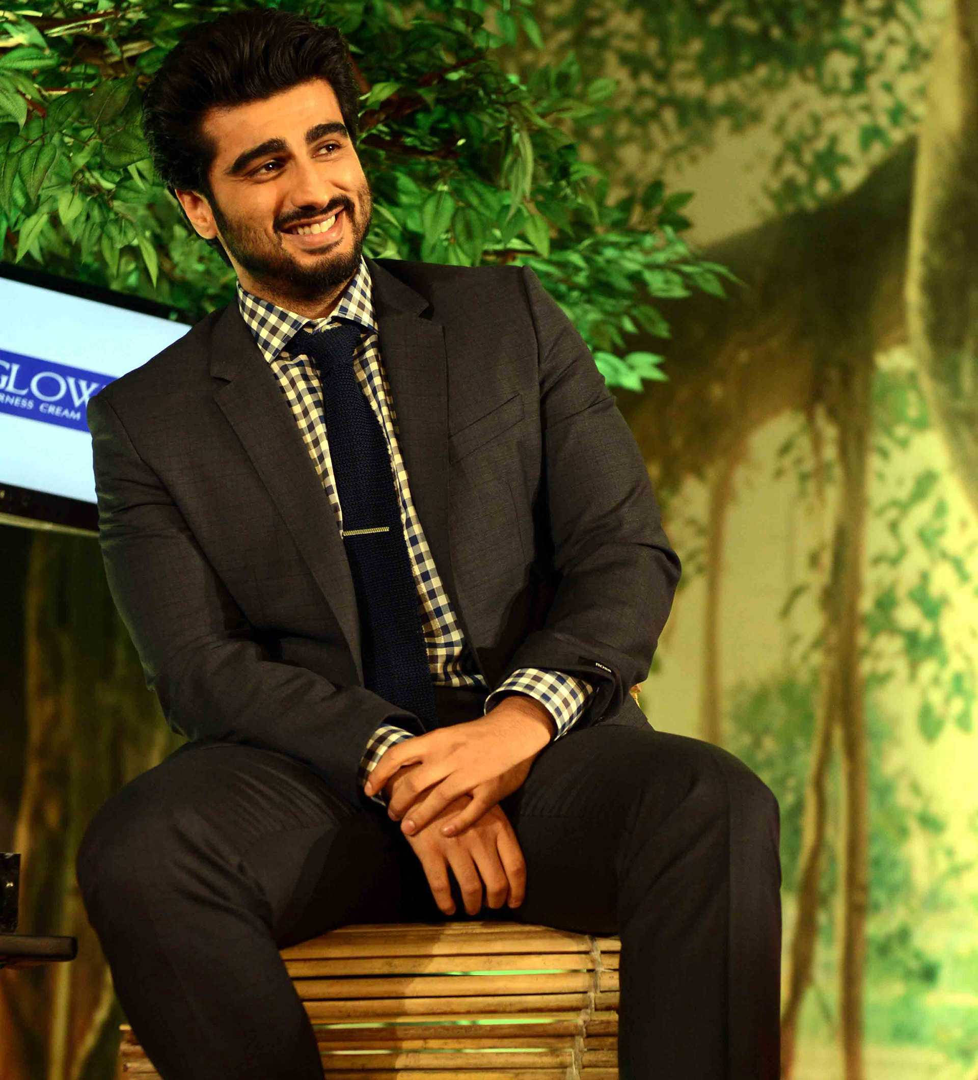 Arjun Kapoor 桌布 壁紙 — 一位微笑的男子身穿深色西裝和格紋襯衫，隨意地坐在竹凳上，背景是茂密的綠色樹葉。